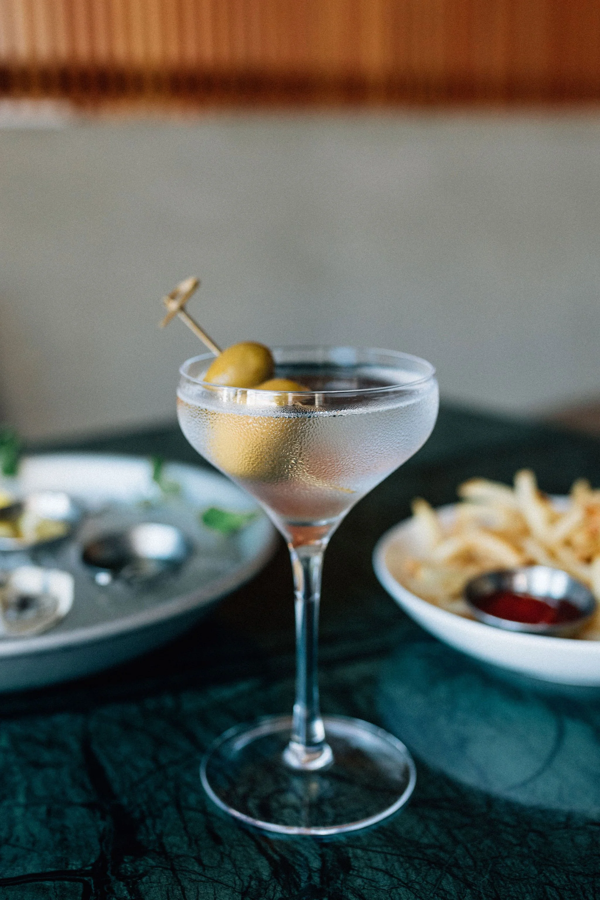 A close-up of a classic cocktail glass filled with a clear cocktail, garnished with green olives on a toothpick, with condensation on the outside of the glass. In the background, there are plates of snacks including potato chips, ketchup, some green 