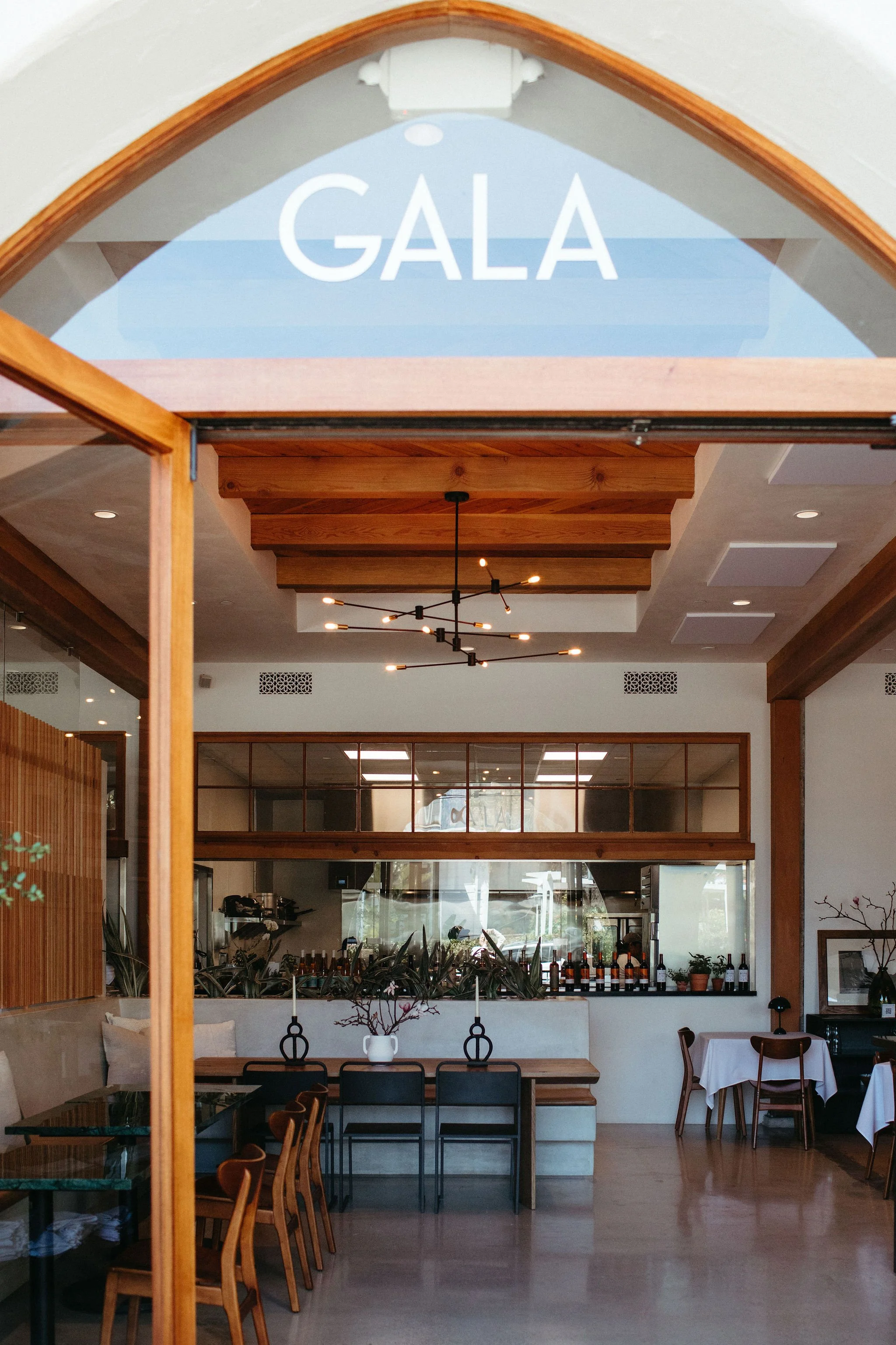 Interior of a modern restaurant with wooden accents, a high ceiling with wooden beams, and a sign that says 'GALA' hanging above. The space has tables with chairs, some covered with white tablecloths, and plants near the counter area.