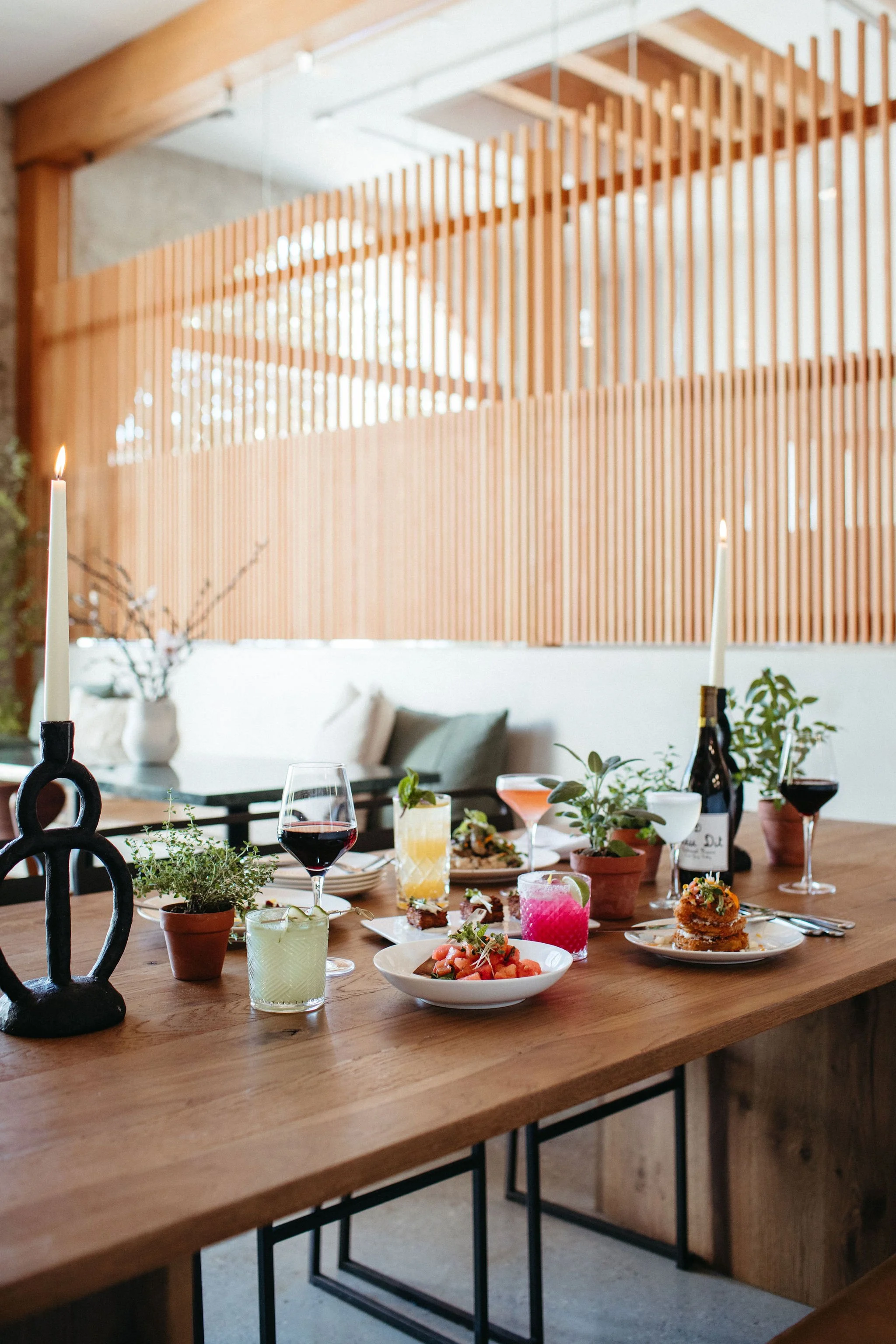 A dining table with various plates of food and drinks, decorated with small potted plants, in a cozy room with wooden accents and a partition wall.