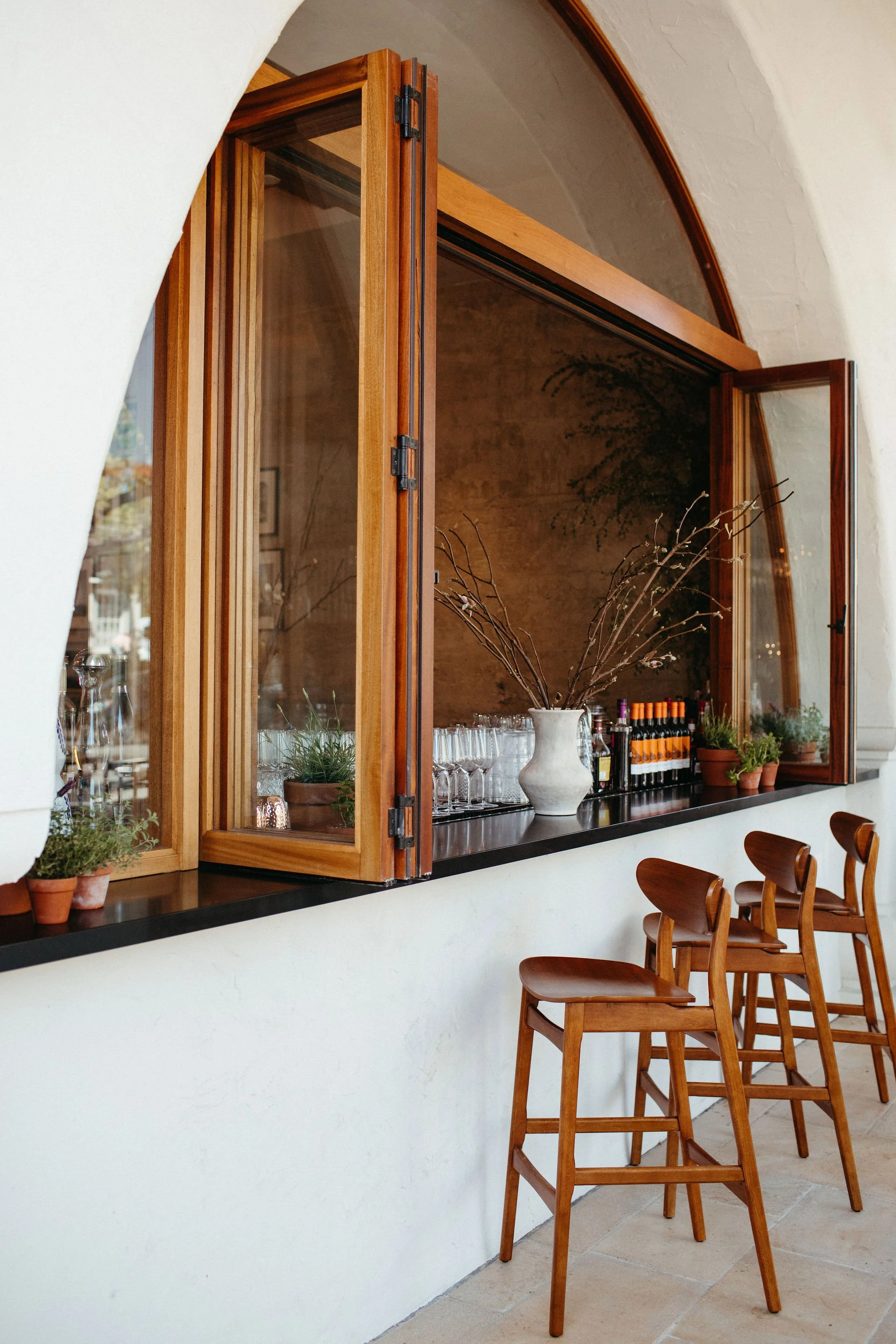 Interior view of a well-lit room with large wooden-framed windows, a white countertop beneath the windows with plants, glassware, and wine bottles, and wooden chairs lined up along the wall.