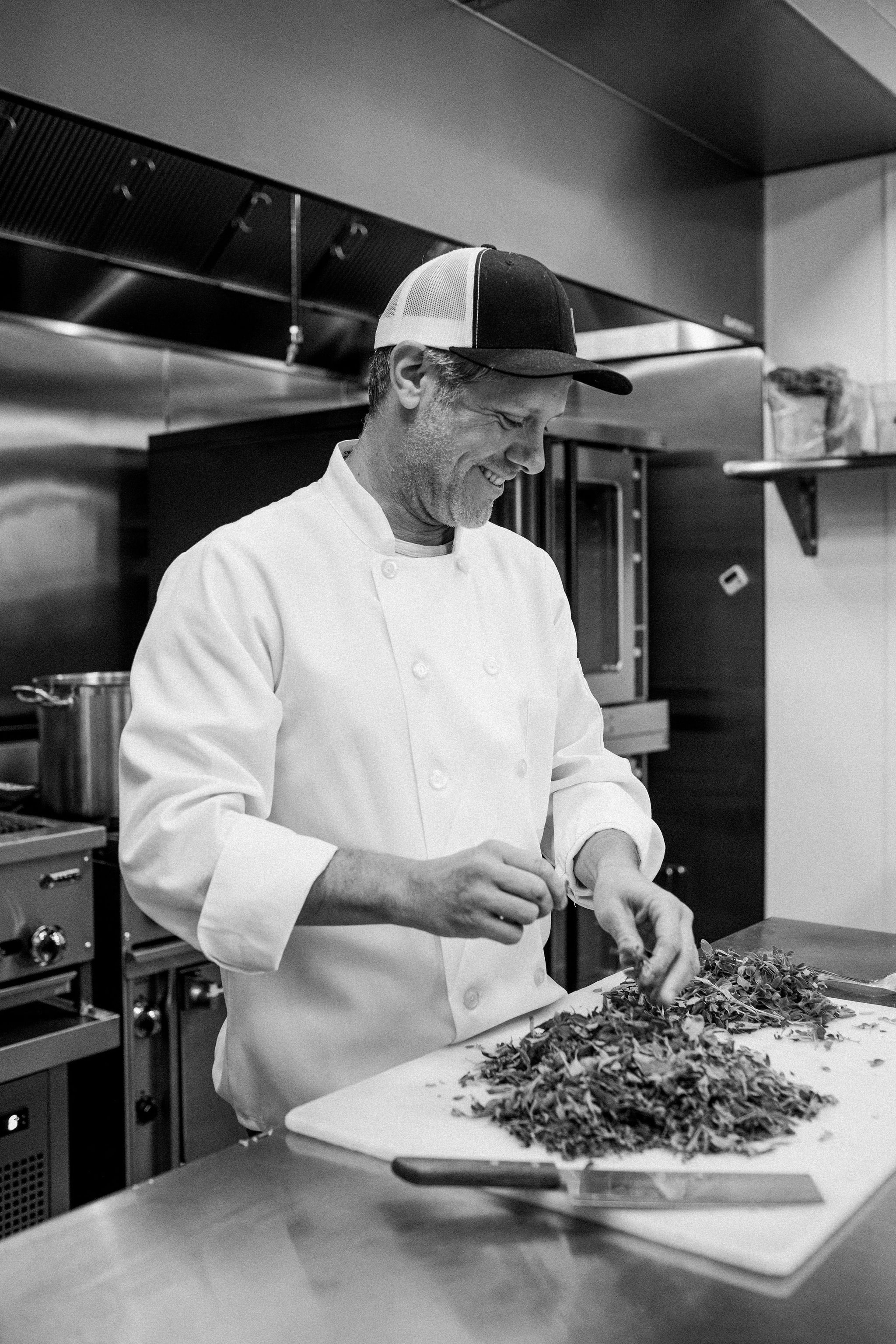 Black and white image of a chef in a professional kitchen, chopping herbs on a cutting board, with piles of chopped herbs in front of him.