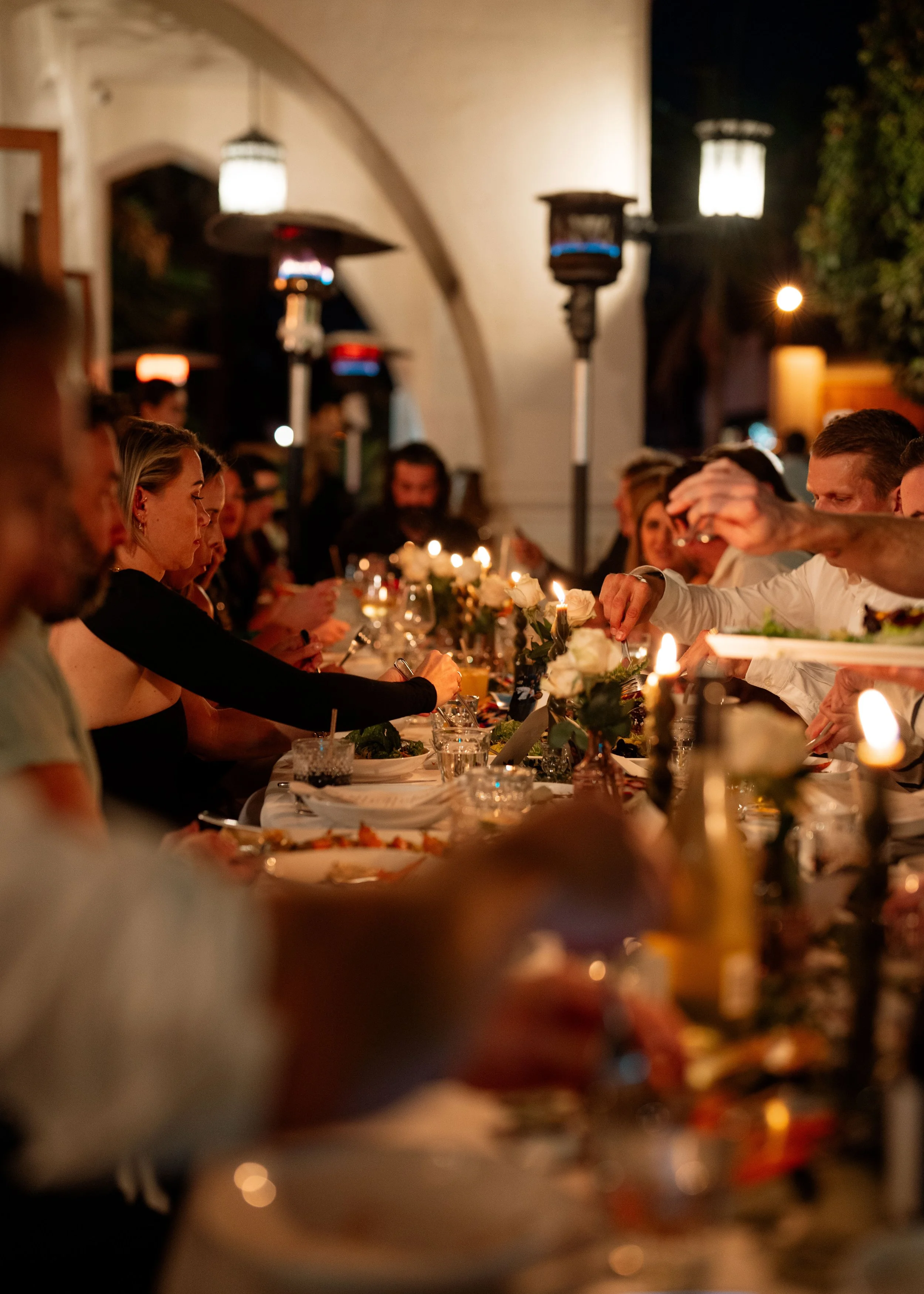 People sitting at a long dinner table during a nighttime outdoor gathering, with lit candles and tea lights, decorative flowers, and string lights outside a building with hanging lanterns.