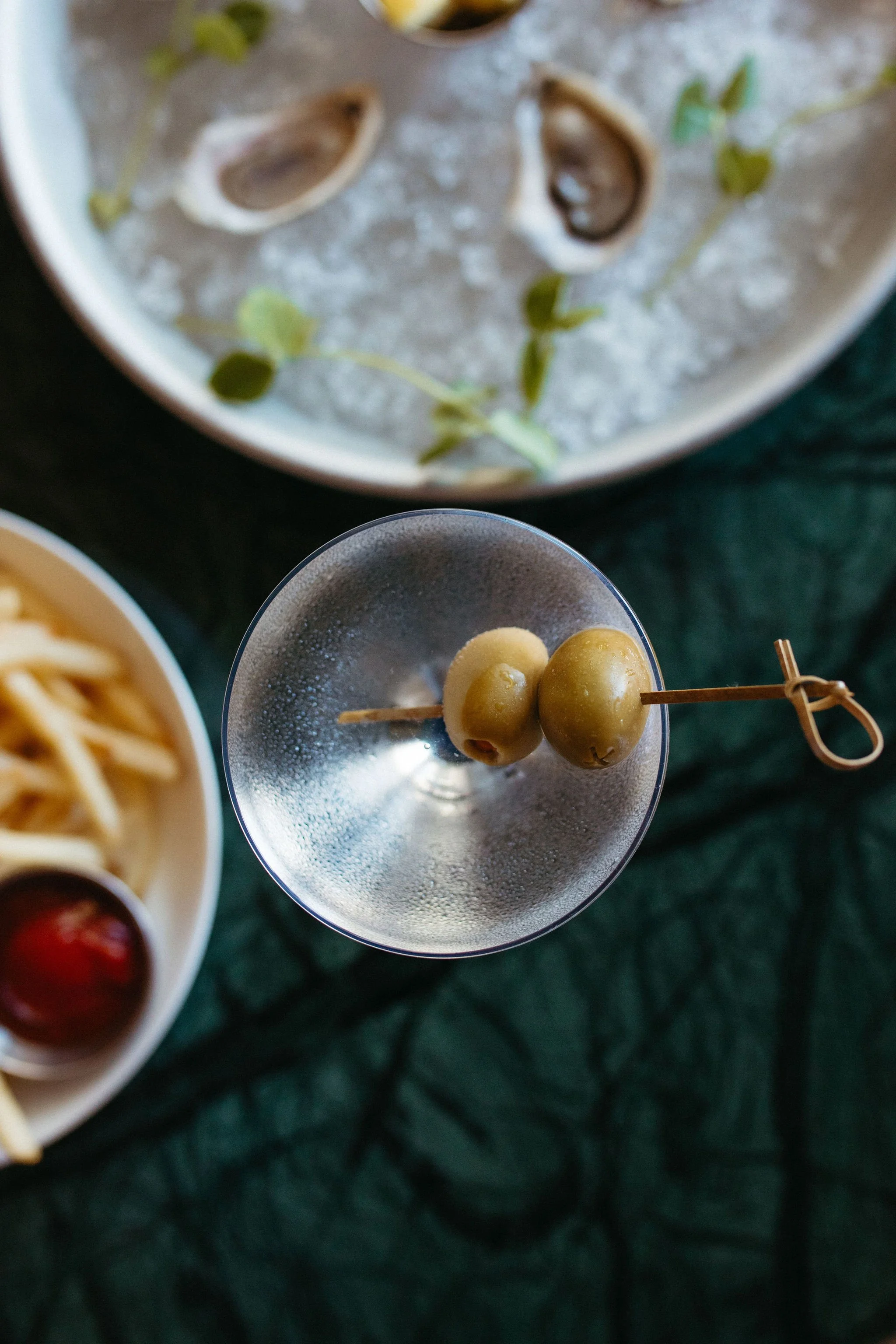 A close-up of three green olives on a skewered toothpick inside a metallic cup, with a blurred background of other dishes and a dark green tablecloth.