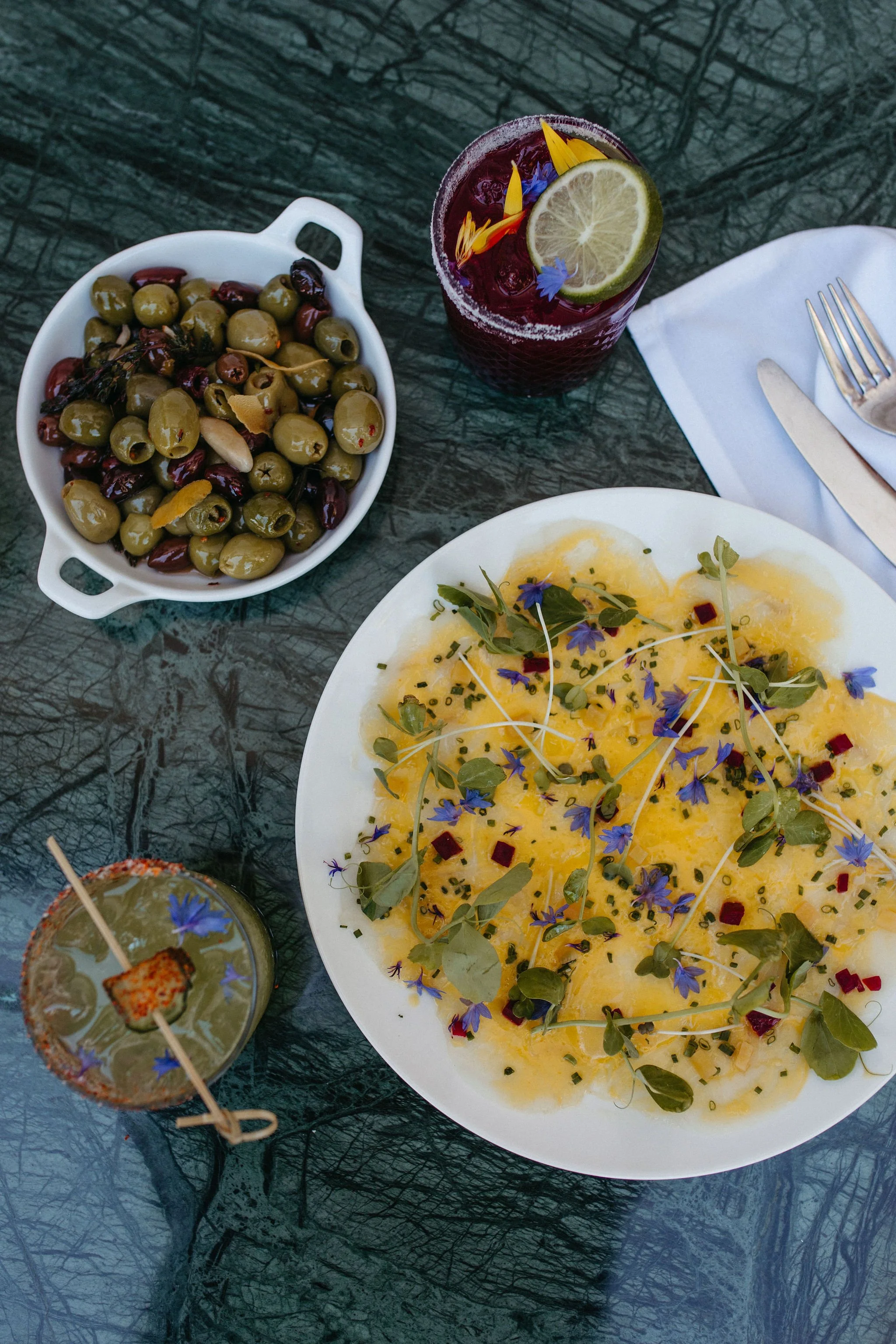 A table set with a bowl of mixed olives, a dessert with edible flowers and microgreens, and a drink garnished with lemon, edible flowers, and blue butterfly pea flowers.