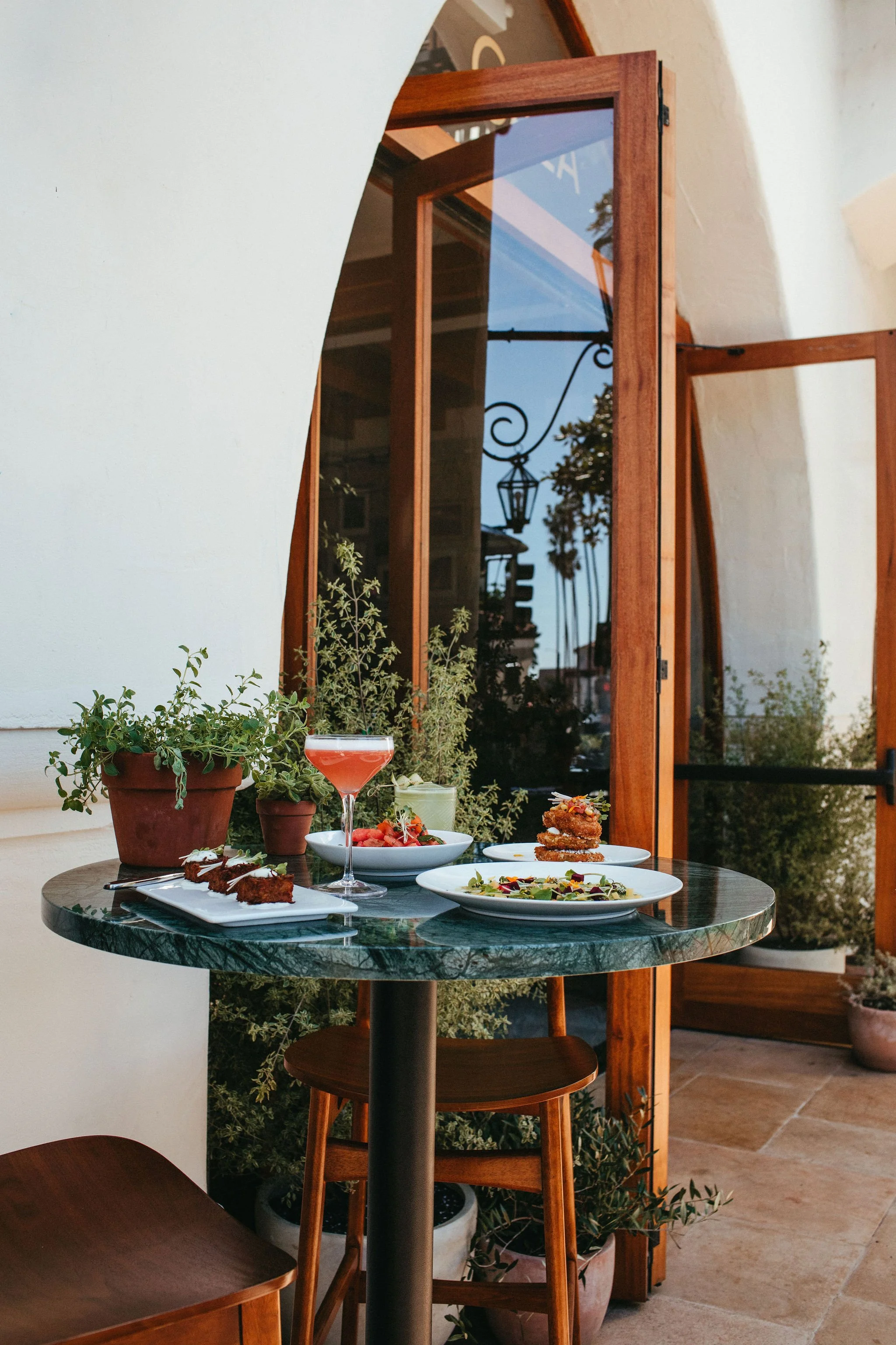 A round marble table set with various dishes including salads, fried chicken, and drinks, surrounded by wooden chairs, with large potted plants, and an open wooden window showing a view of trees and a street lamp outside.