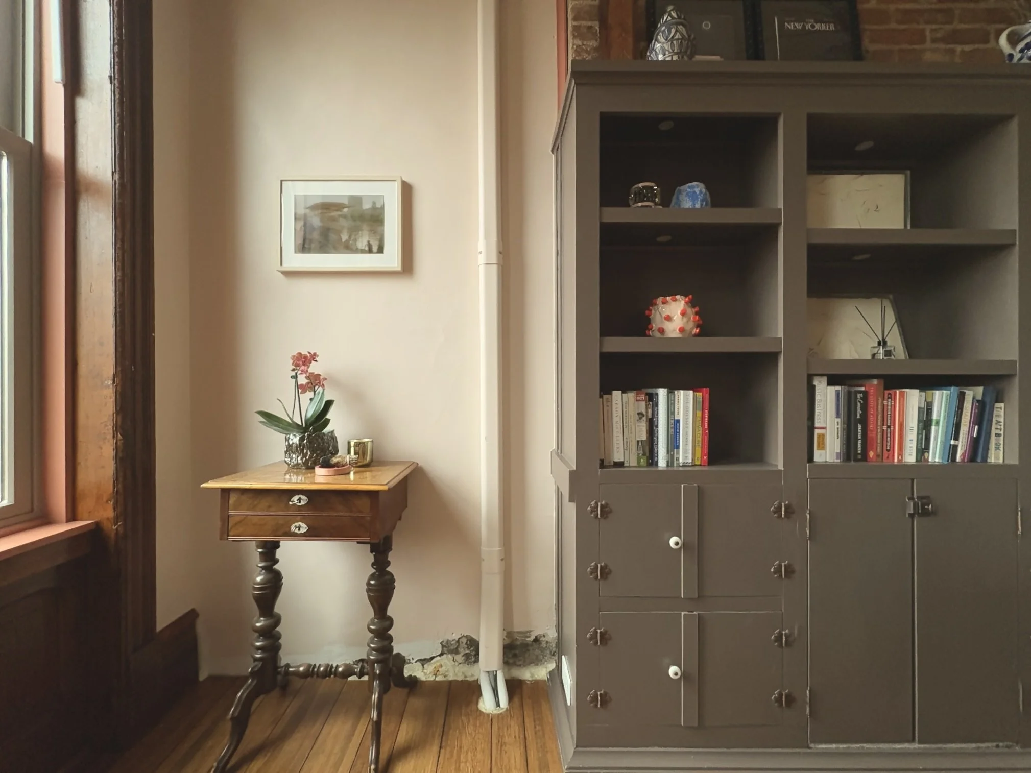 A corner of a room with a small wooden table holding a potted plant and a cup, a framed picture on the wall, a gray bookshelf with books and decorative items, and a window with a wooden trim.