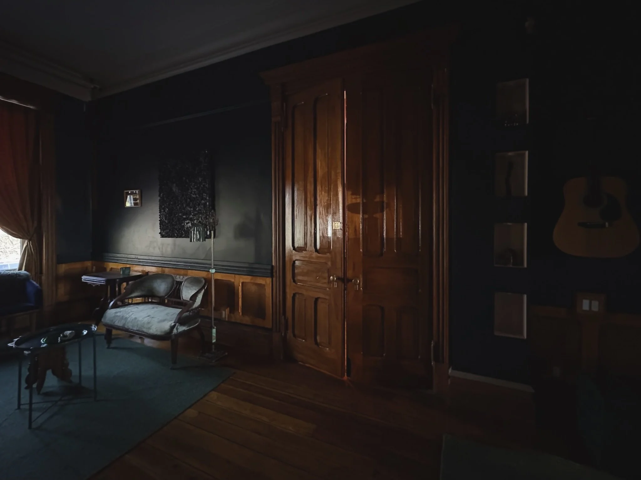 Living room with wooden flooring, dark blue walls, wooden door, and furniture including a vintage sofa, wall art, and guitar