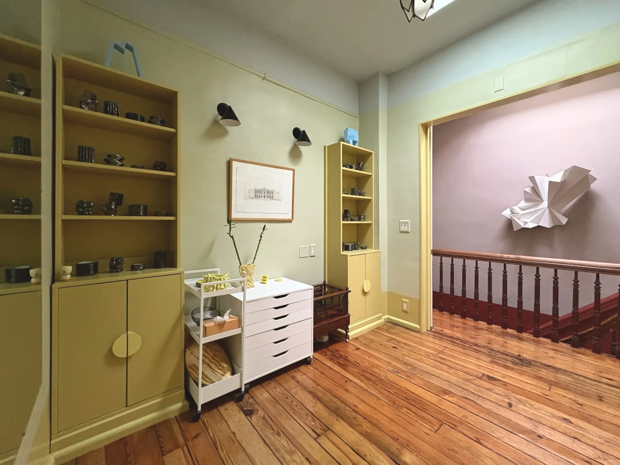 Interior view of a room with wooden floors, yellow and beige walls, and bookshelves, including a corner with a railing and wall art.