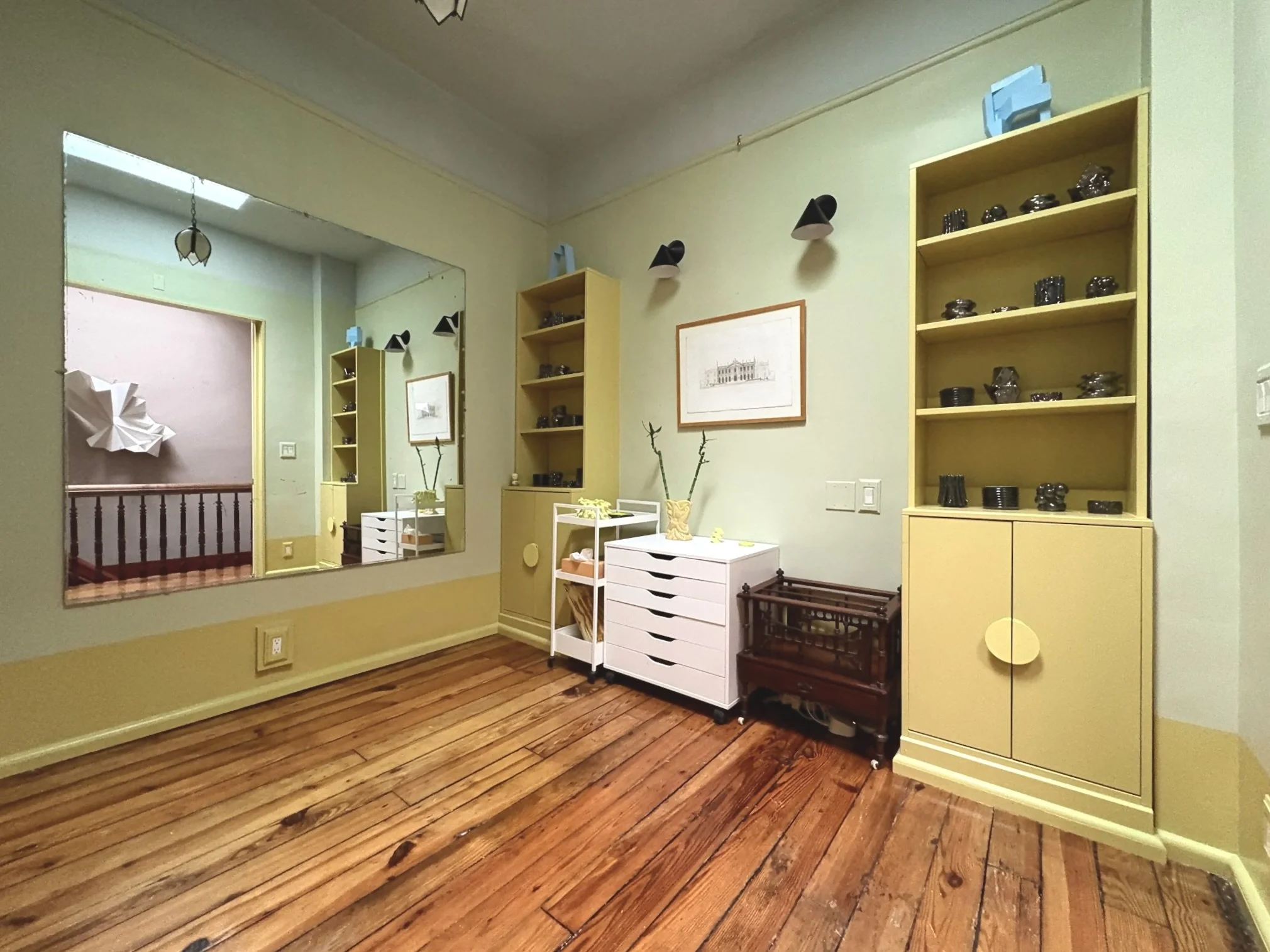 Interior view of a room with light yellow walls and wooden flooring, featuring a large mirror, two yellow bookshelves, a white storage cart, a small dark wooden crib, a framed picture on the wall, and various decorative objects.