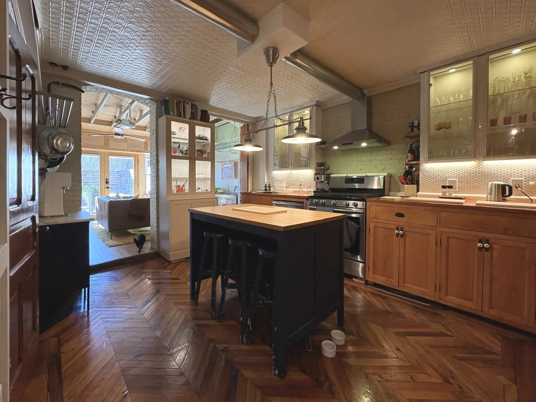Kitchen with wooden cabinets, stainless steel stove, black island with wooden top, pendant lights, and a view into a living room area.