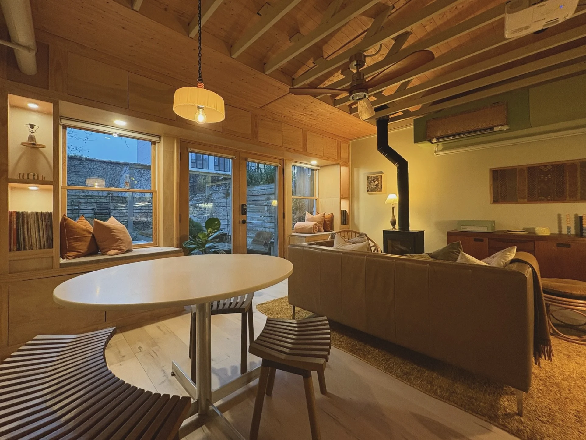 Cozy living room with wooden walls and ceiling, large windows, and a wood-burning stove. Contains a round dining table with chairs, a leather sofa, a side chair near the window, a bookshelf, and ambient lighting.