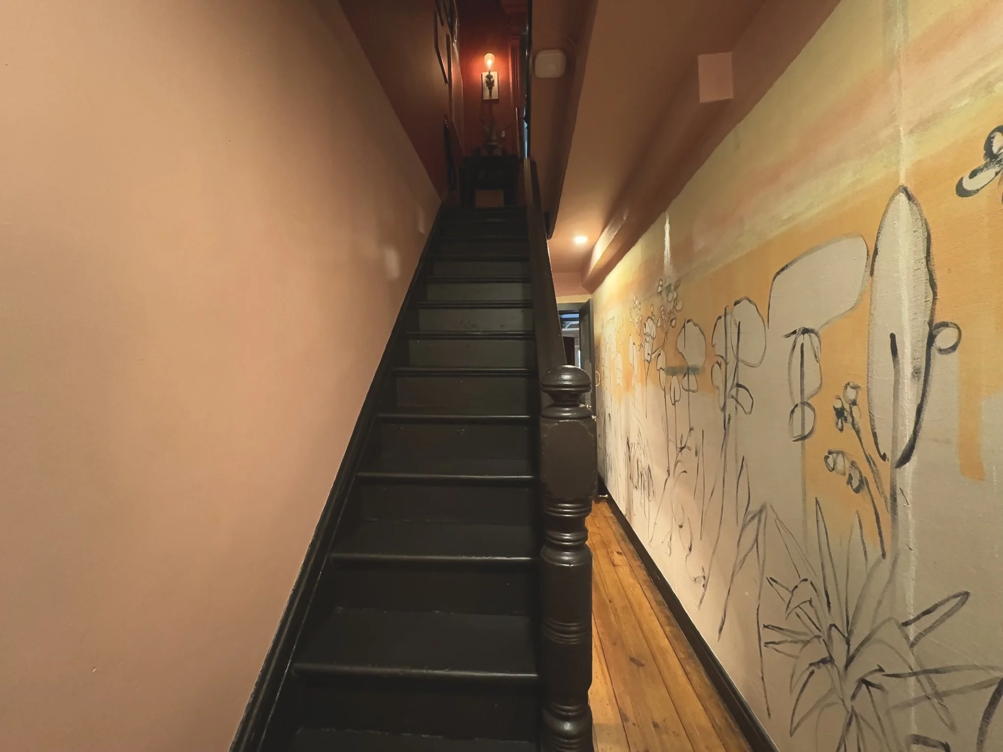 Interior hallway with wooden stairs and a wall mural of flowers painted in soft colors, illuminated by wall-mounted lights.