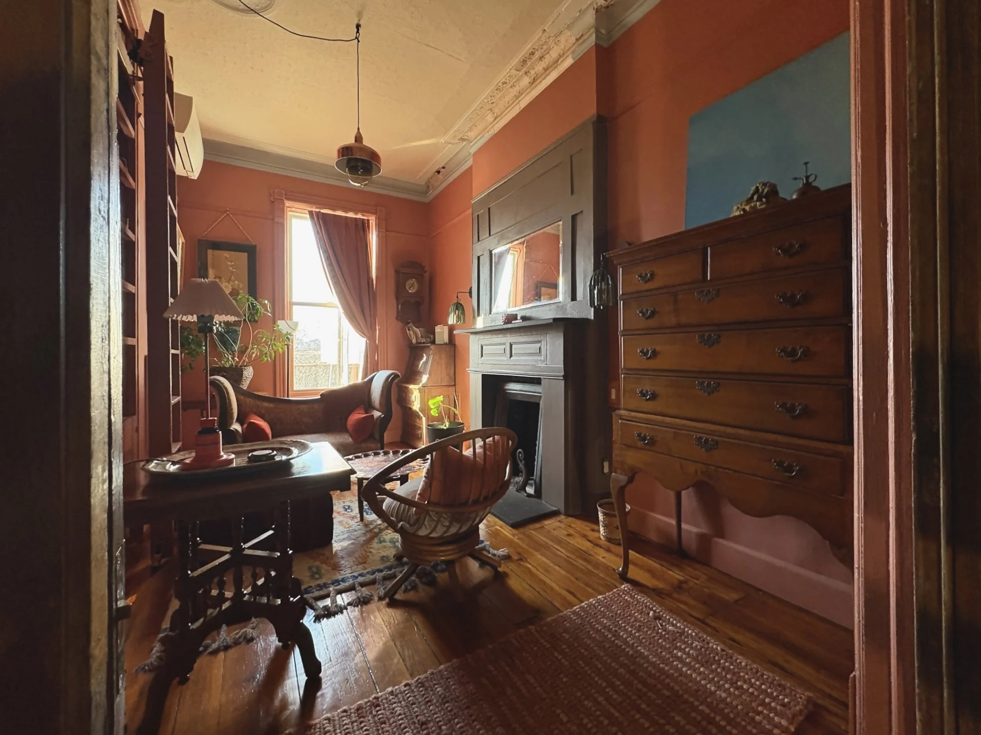 A cozy living room with pink walls, wooden flooring, and antique furniture. There's a window with sunlight, a leather armchair, a small table with a lamp, a wooden chest of drawers, and a fireplace with a mirror above it. The room is decorated with p