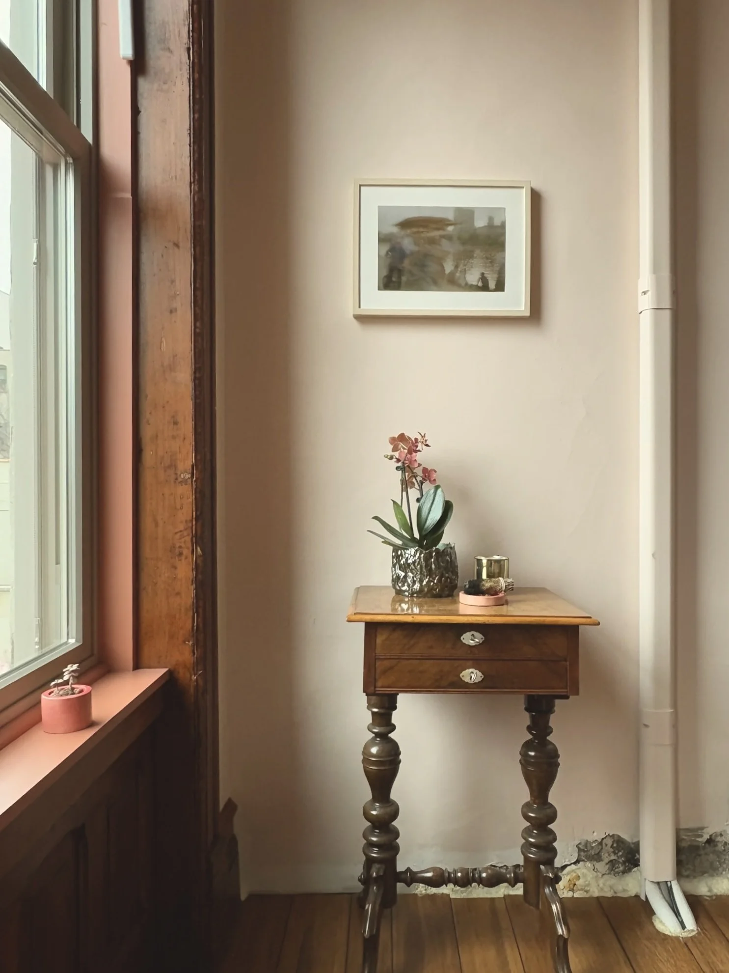 Interior of a cozy corner with a small wooden table holding a potted orchid, candles, and decorative items. A window with a pink window sill and a small plant, framed artwork on the wall, and a exposed pipe at the bottom of the wall.