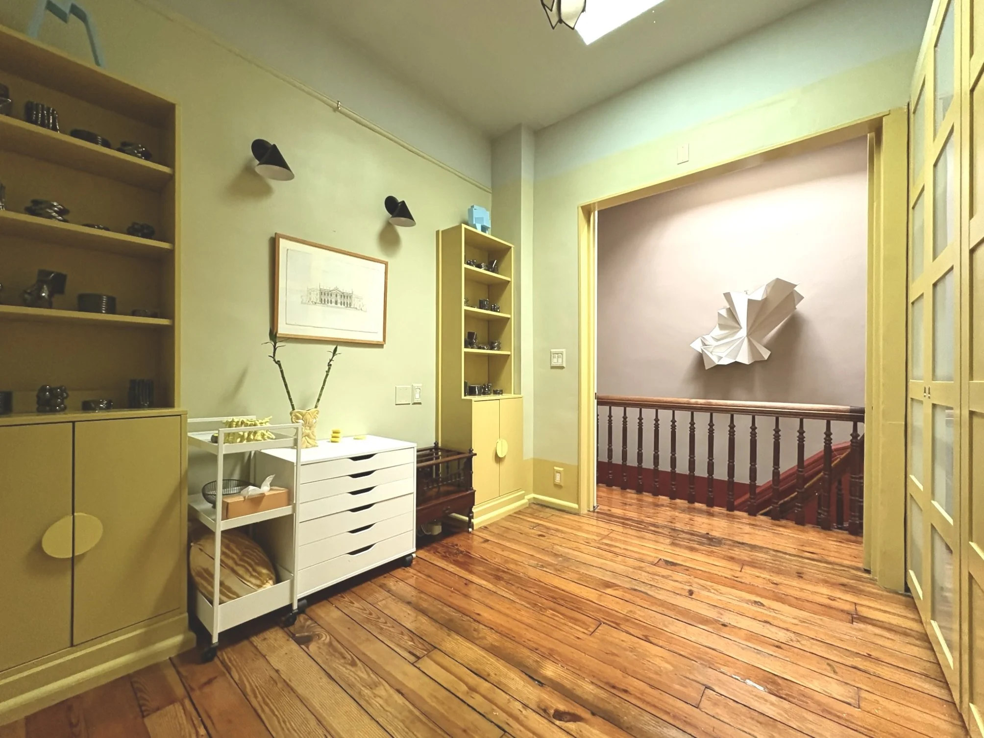 Interior of a room with green walls, wooden floors, yellow shelving units, and a small white trolley with drawers. Decor includes framed artwork, wall-mounted lights, and abstract wall art on the stairwell landing.