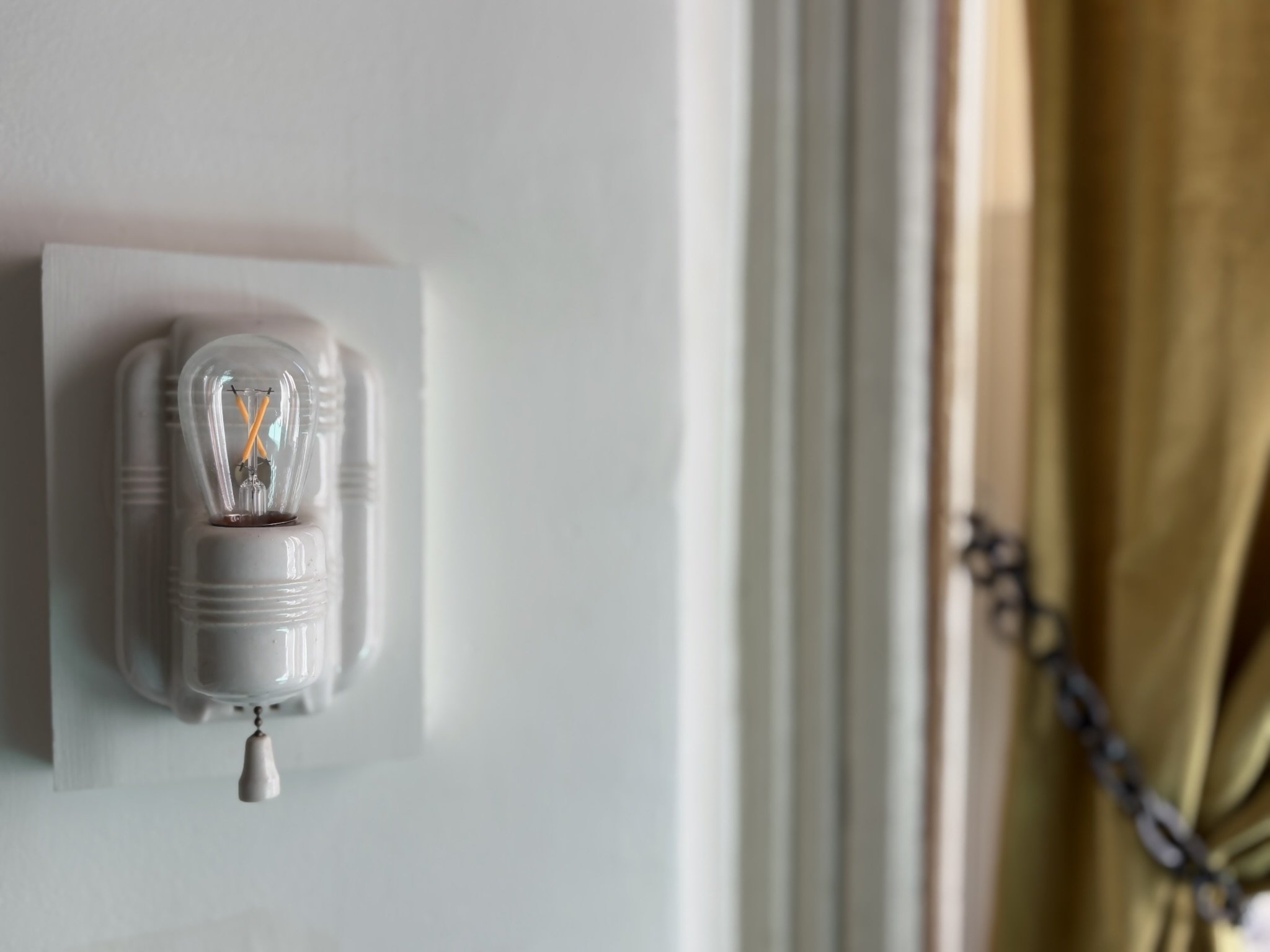 A close-up of a small light bulb with a decorative filament mounted on a white wall, with a patterned curtain in the background.