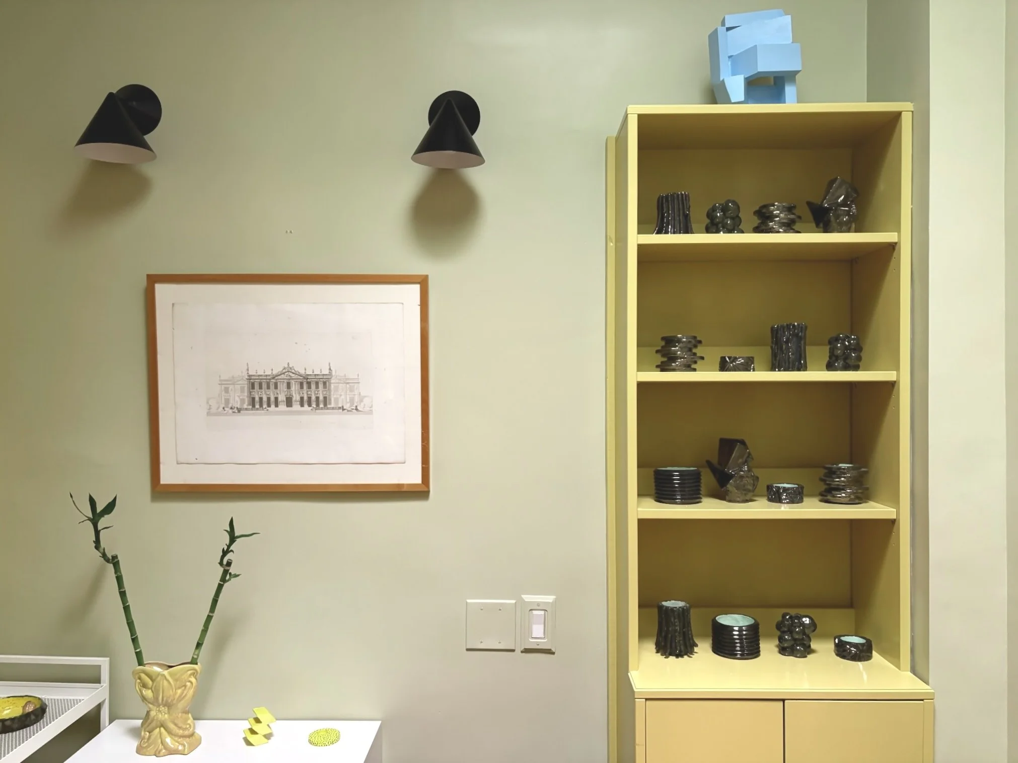 Interior scene with a framed drawing of a classical building on a light green wall, two black wall-mounted lights, a tall yellow bookshelf with black and metallic decorative objects, a yellow ceramic vase with green bamboo stalks on a white table, an