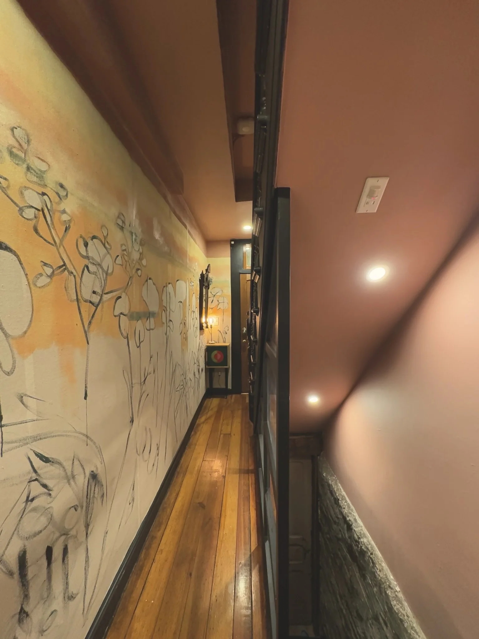 Residential interior hallway with painted floral mural on the left wall, polished wooden floor, and soft overhead lighting. There is a small table with a lamp at the far end.