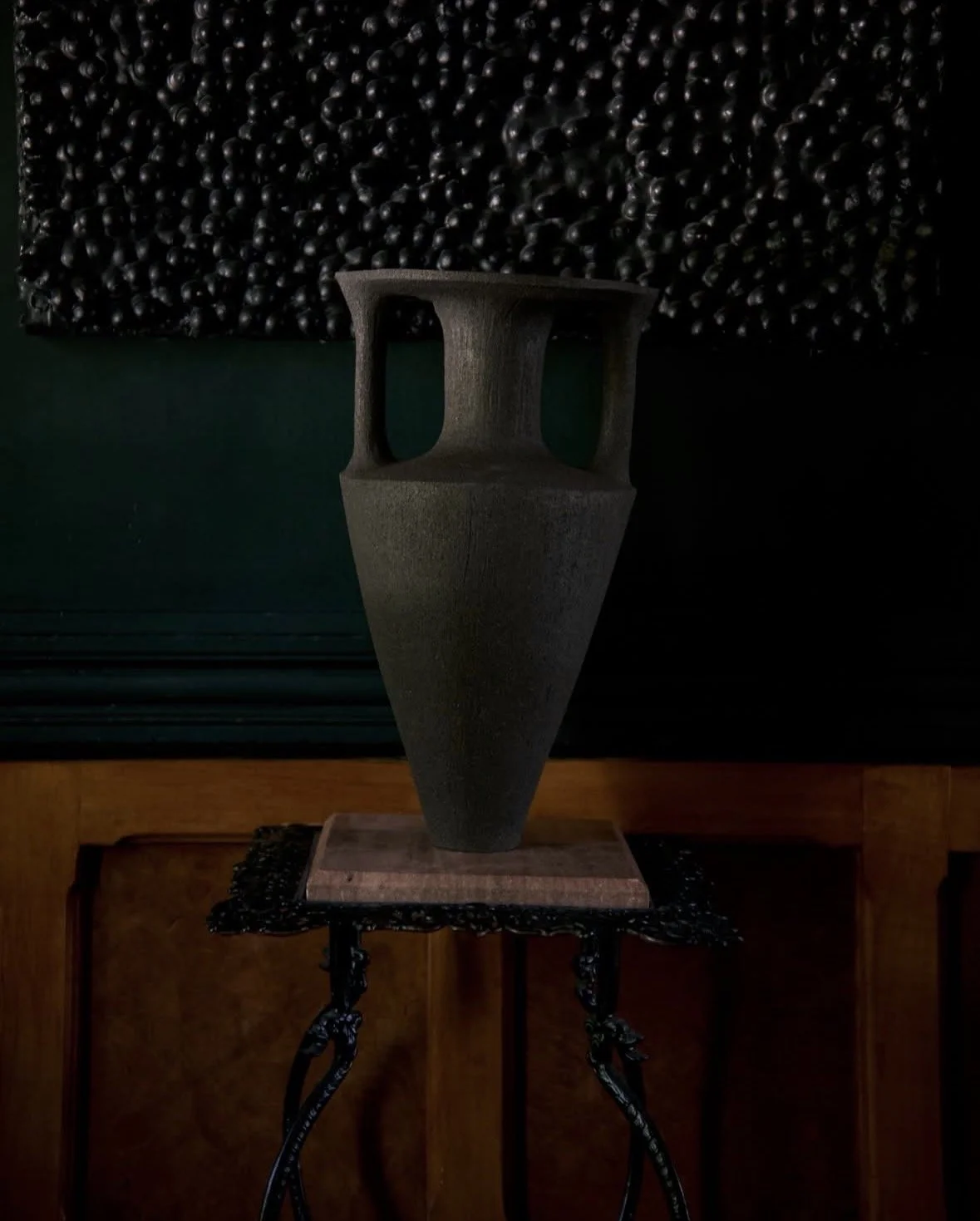 A tall, narrow dark-colored vase with two handles on a wooden platform, placed on an ornate black metal stand, with a wall of dark berries and green panels in the background.