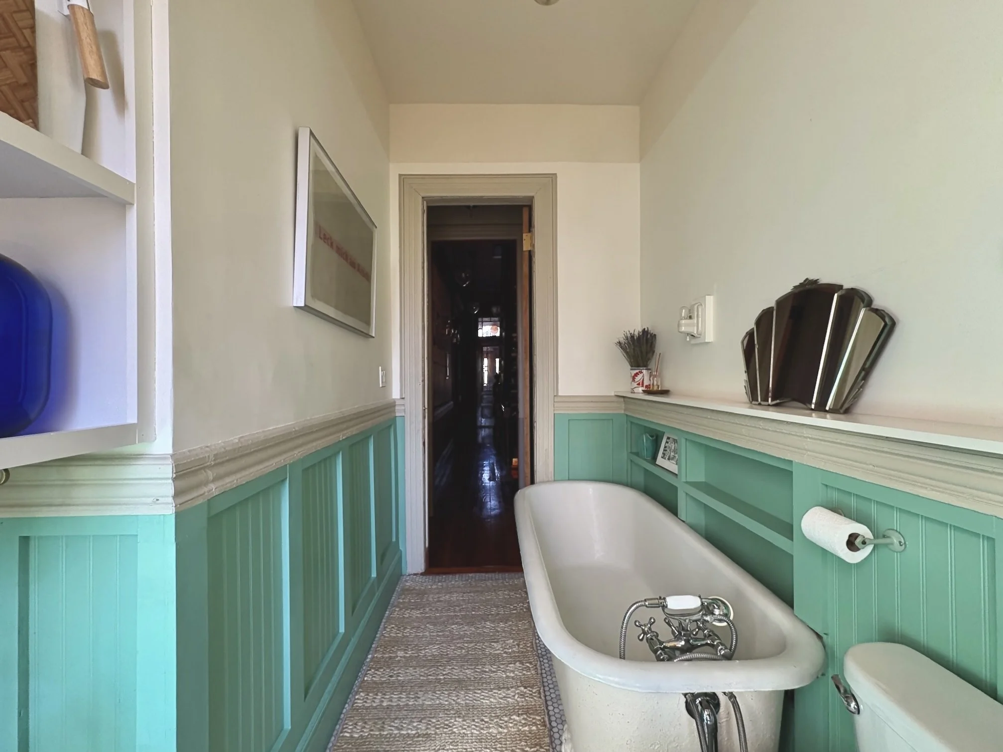 A bathroom features an oval clawfoot bathtub, green beadboard wainscoting, and wall-mounted mirrors. There is a wicker basket with dried lavender and a roll of toilet paper on the wall.
