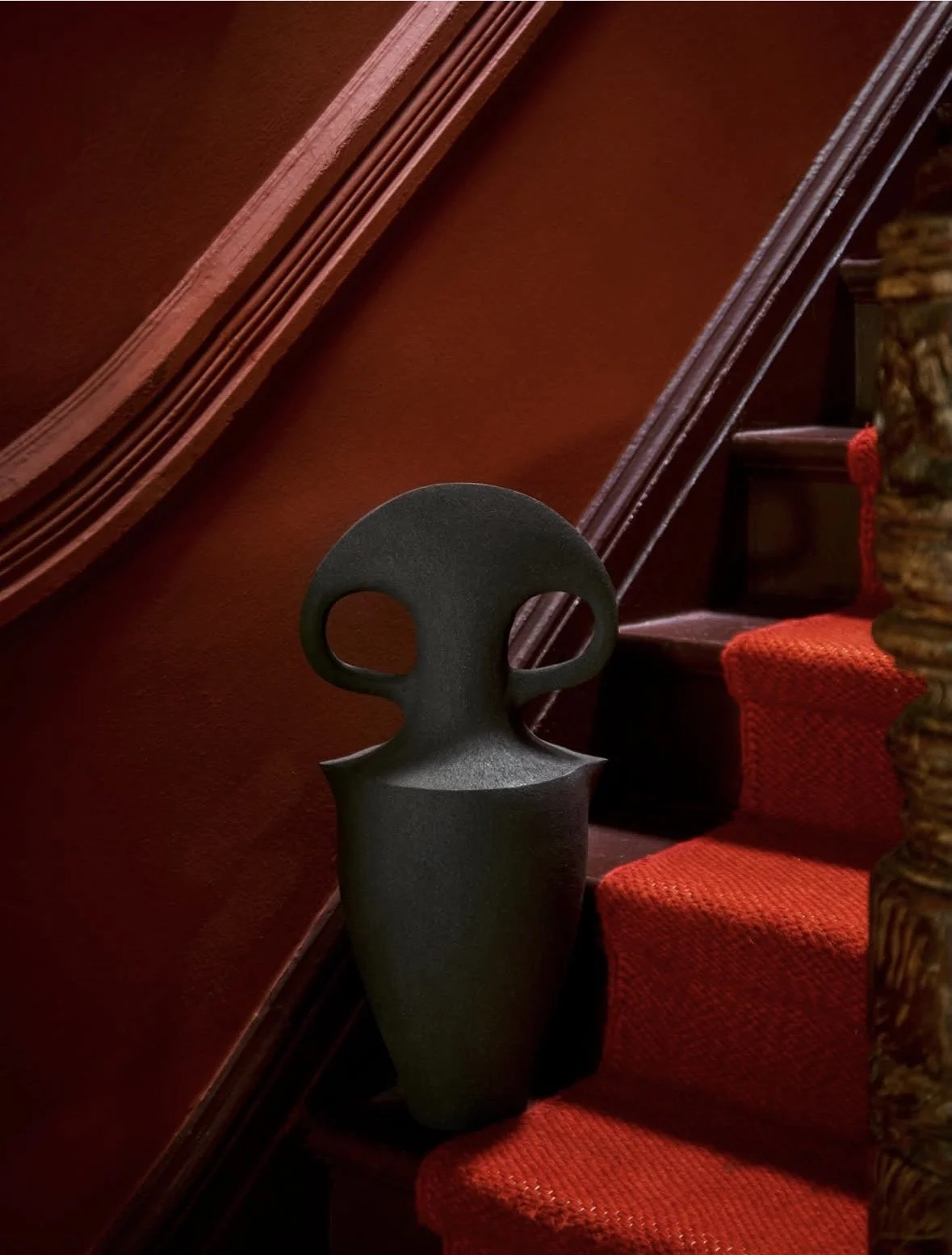 A black modern decorative sculpture resembling a face with two large eye holes, placed on a staircase with red carpeting and wooden handrails.