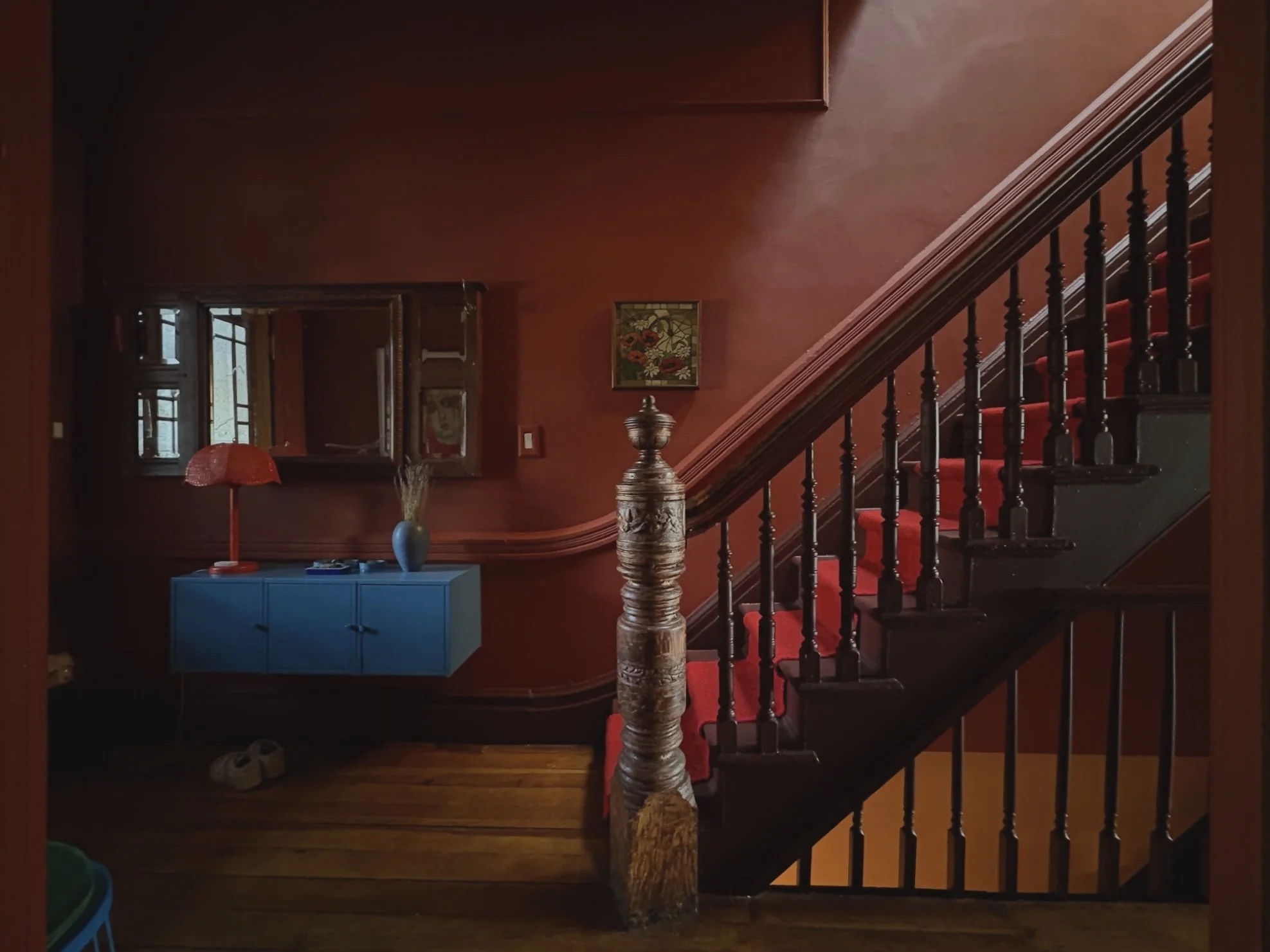Interior of a home staircase area with a red wall, a blue sideboard, a orange table lamp, a blue vase with dried flowers, and a window reflection showing outside scenery.