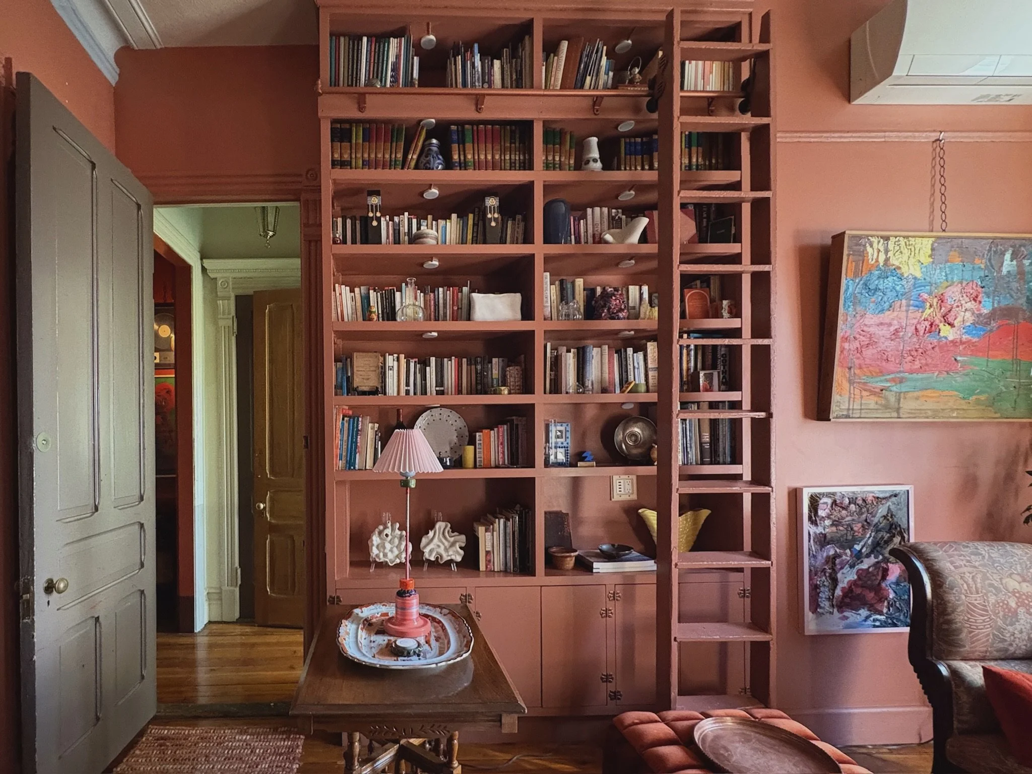 A room with pink walls featuring a large pink wooden bookshelf filled with books, decorative objects, and photographs. To the right, there is a colorful abstract painting and a smaller artwork below it. A wooden table with a pink table lamp and tray 