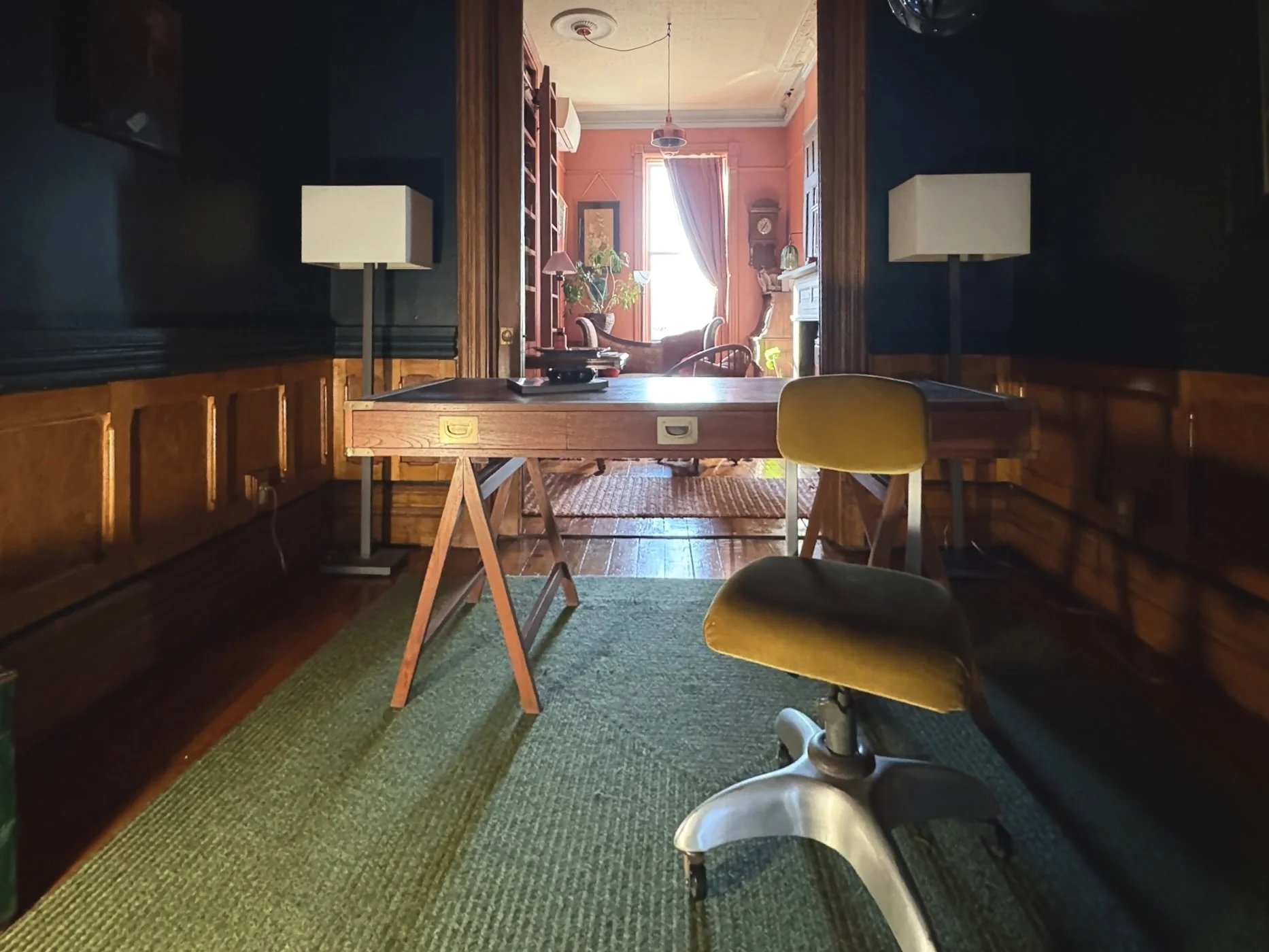 A vintage-style room with dark-paneled walls, a wooden table, yellow chair, and two white floor lamps. In the background, a bright room with pinkish-orange walls, a window with curtains, a potted plant, and an armchair.