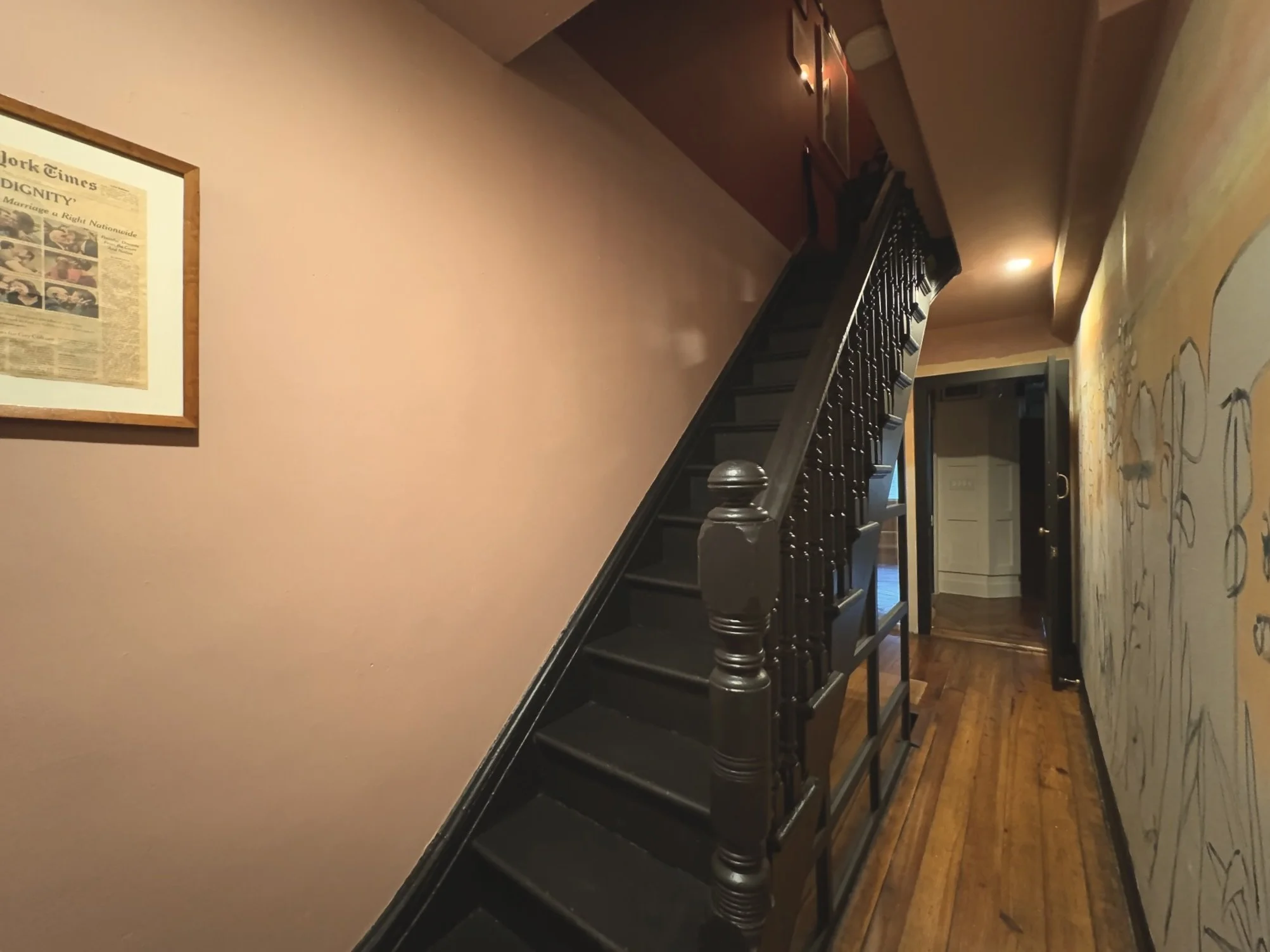 Interior view of a hallway with a staircase, wooden floors, framed newspaper on the wall, and a mural or artwork on the other wall.