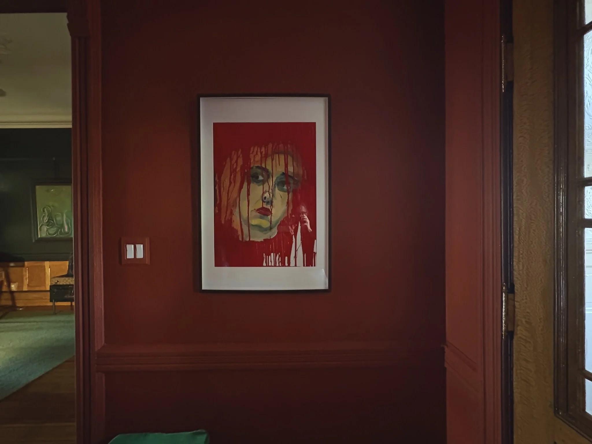 A framed artwork painting of a woman's face with red hair and makeup, hanging on a red wall in a room with wooden trim and a window on the right side.