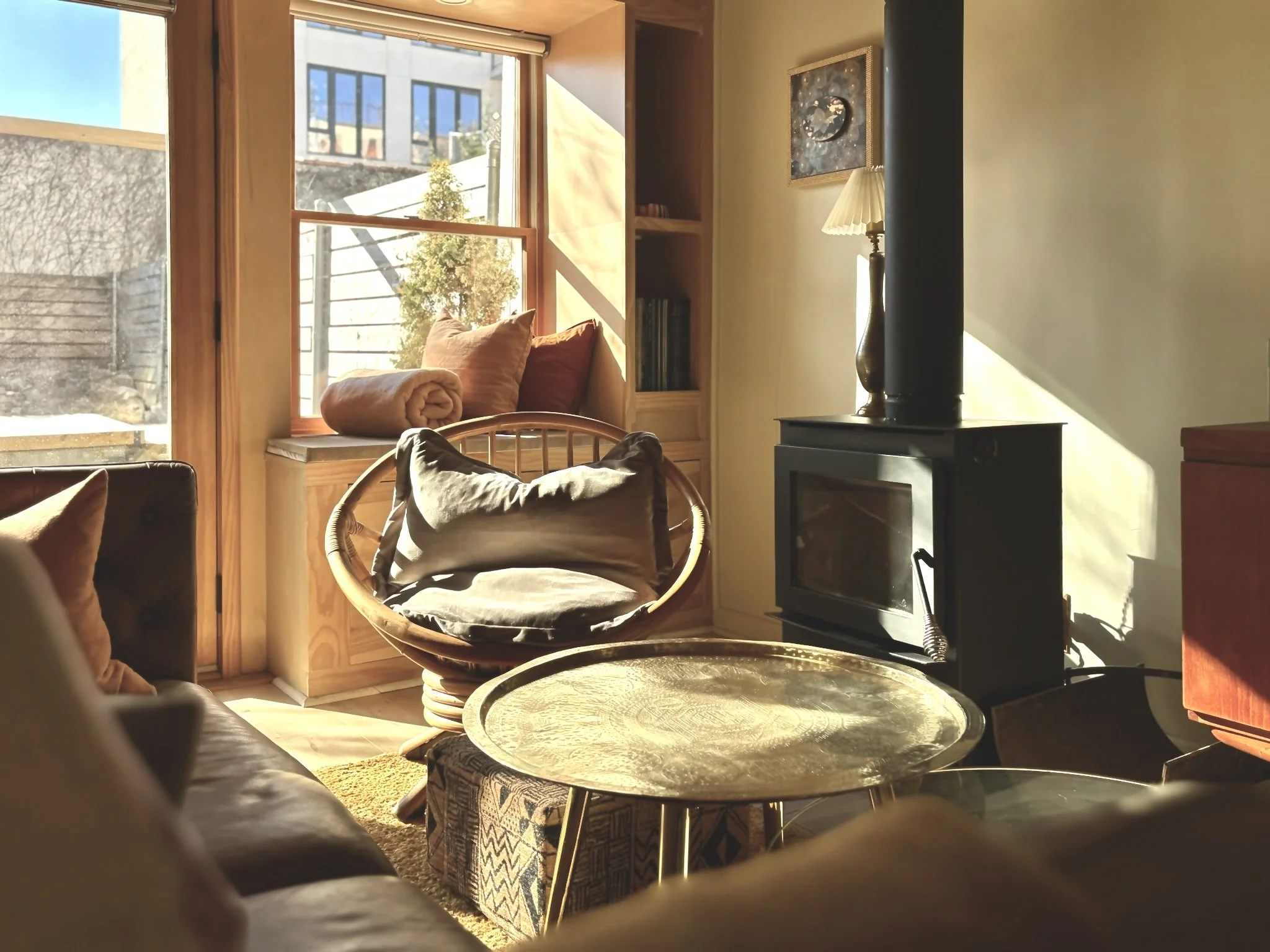 Sunlit cozy living room with a wood stove, a cushioned chair, a window seat with pillows, shelves with books, and a round coffee table.