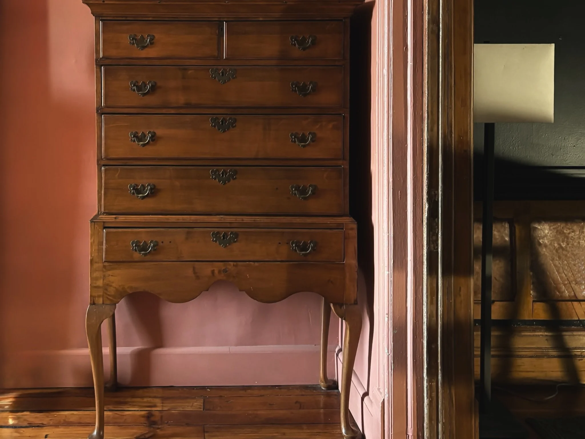 A wooden vintage dresser with five drawers and ornate handles, standing against a pink wall, with a leg of another piece of furniture visible at the bottom.