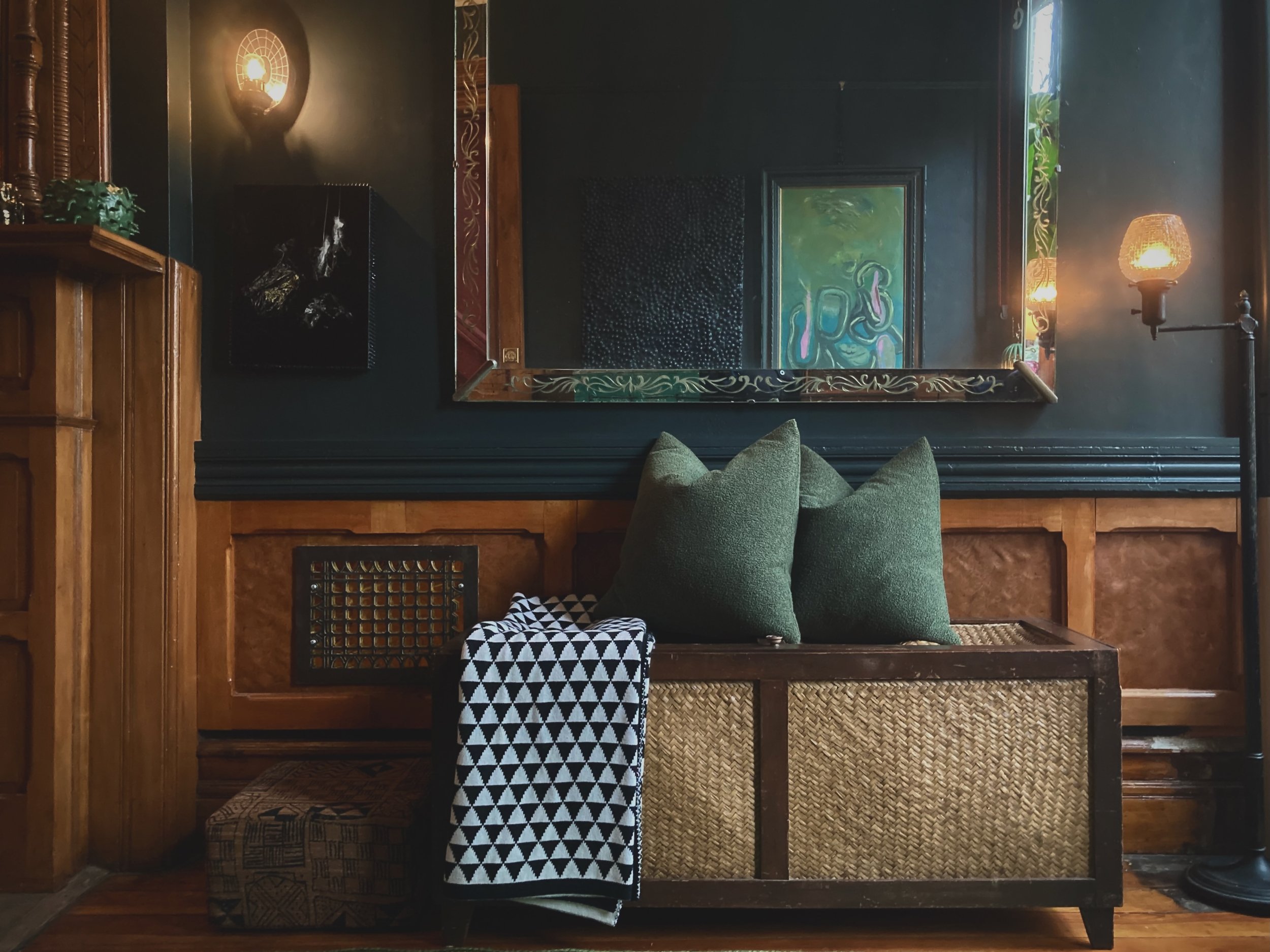 Interior living space with dark walls, featuring a large mirror with decorative border, two wall-mounted light fixtures, a wooden cabinet, a bench with green pillows, a black-and-white patterned blanket, and artwork on the walls.