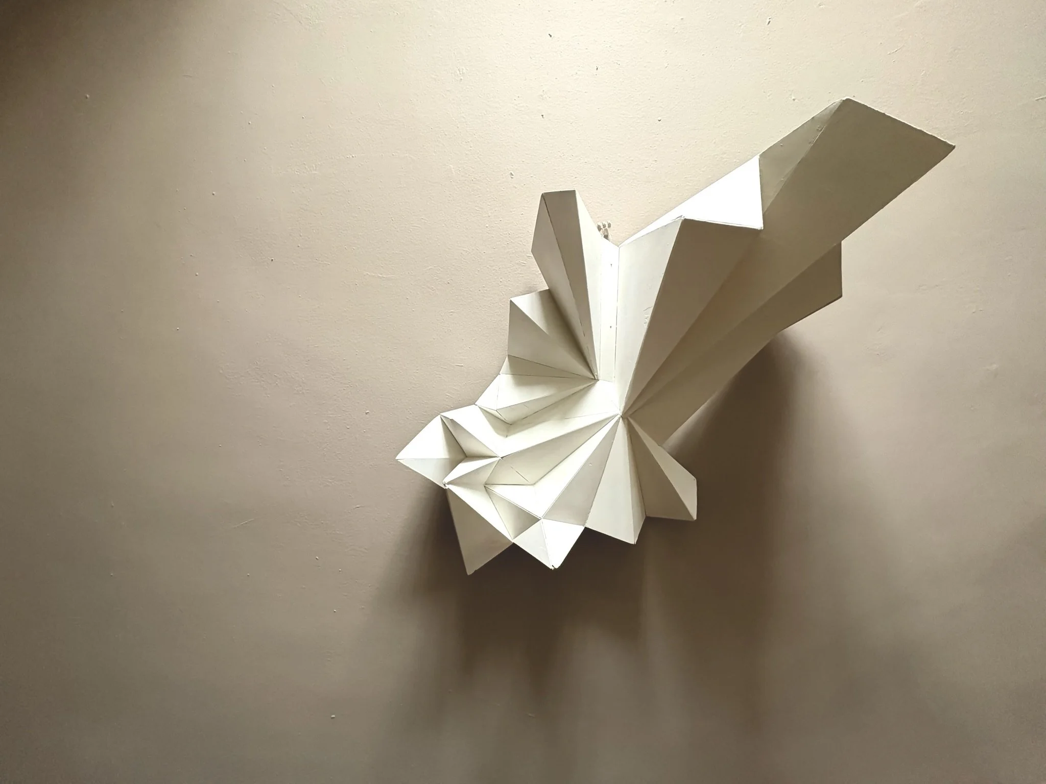 White geometric paper sculpture with folded and angular design, mounted on a beige wall.