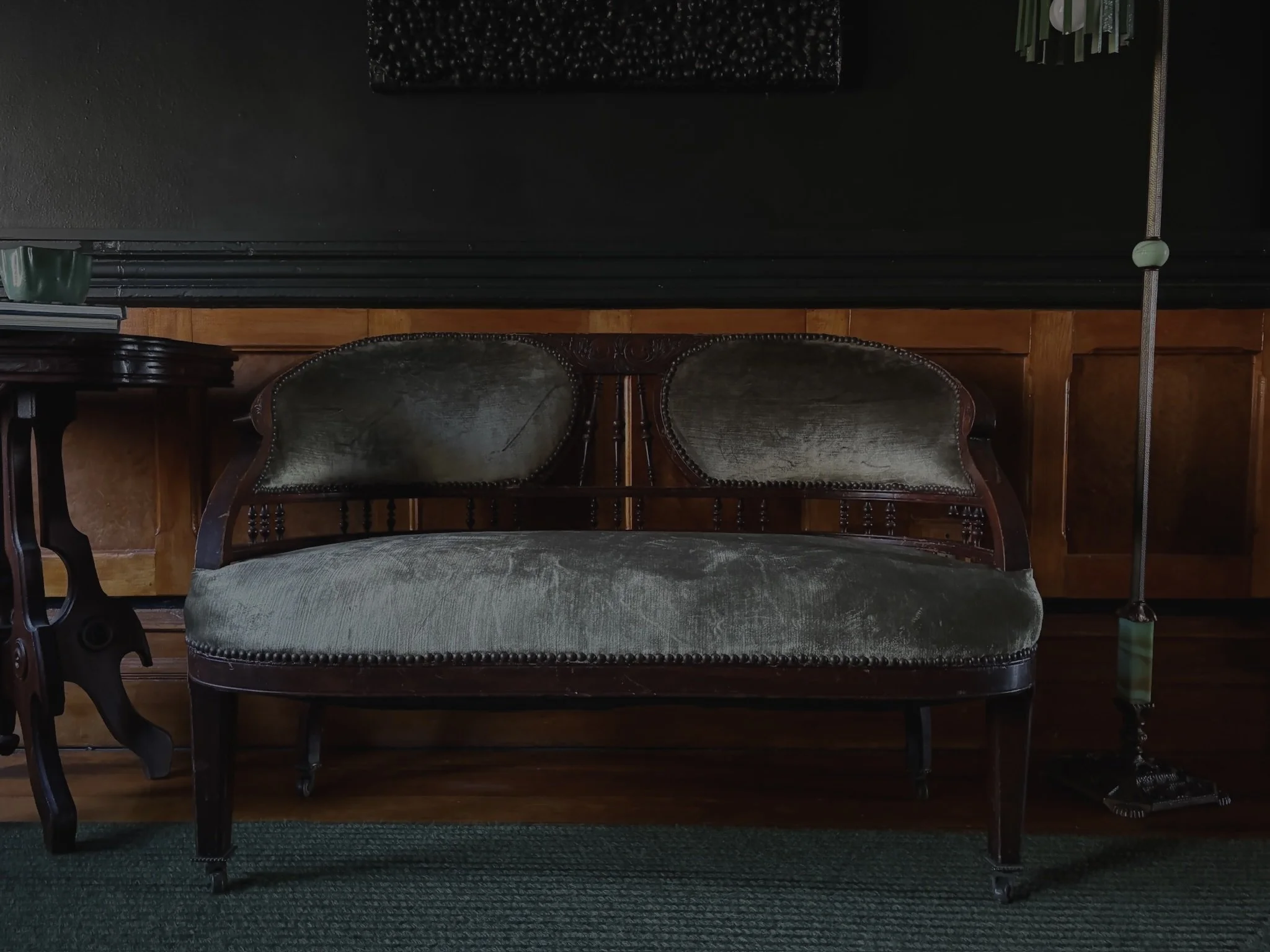 Vintage wooden loveseat with dark green velvet upholstery, positioned against wood-paneled wall, with a floor lamp on the right and a side table on the left.