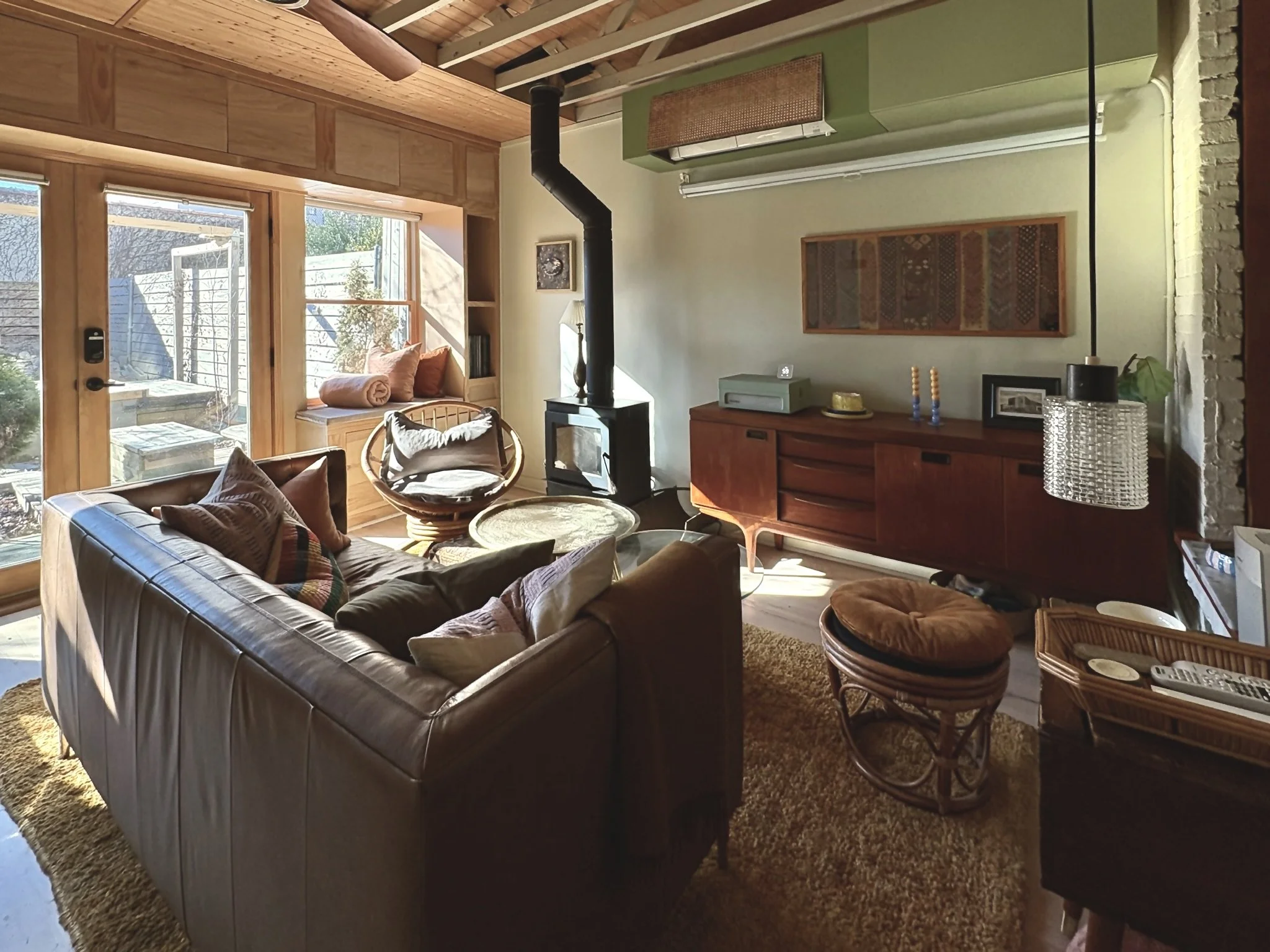Cozy living room with a leather sofa, wicker chairs, a wood stove, and large windows offering sunlight and outdoor views.