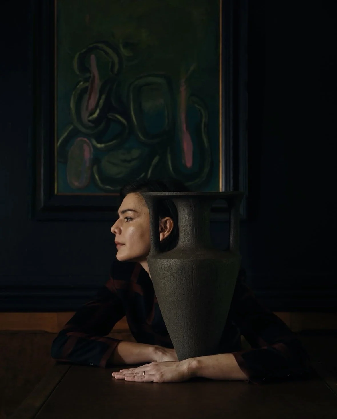 A woman with a uniquely shaped head sculpture sitting at a table in a dark room with a dark abstract painting on the wall behind her.