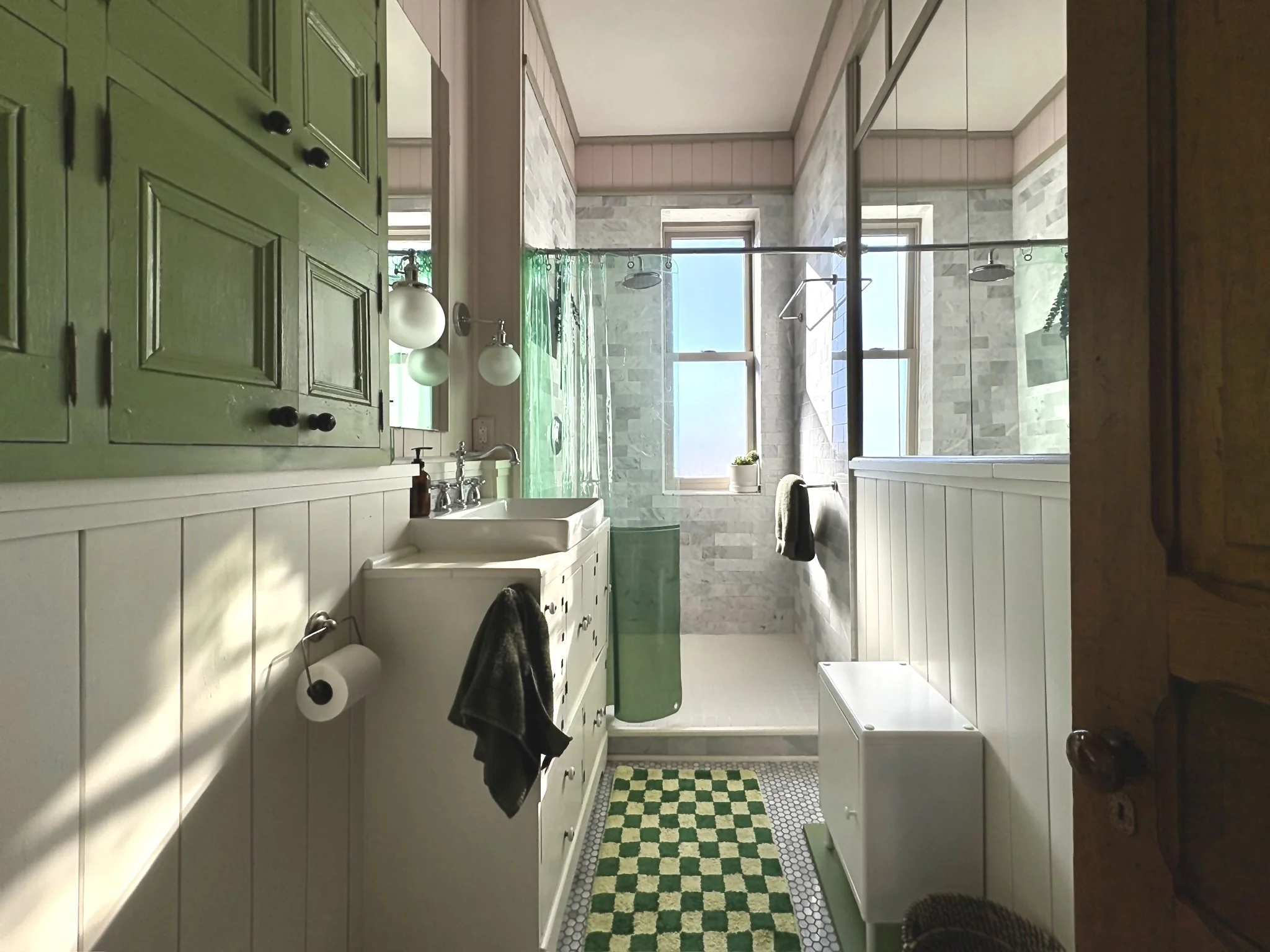 Bright bathroom with a green and white color scheme, featuring a double vanity with a white sink, a wall-mounted mirror, and globe lights. There is a checkered green and yellow rug on white hexagon tiles, a walk-in shower with glass doors, large wind