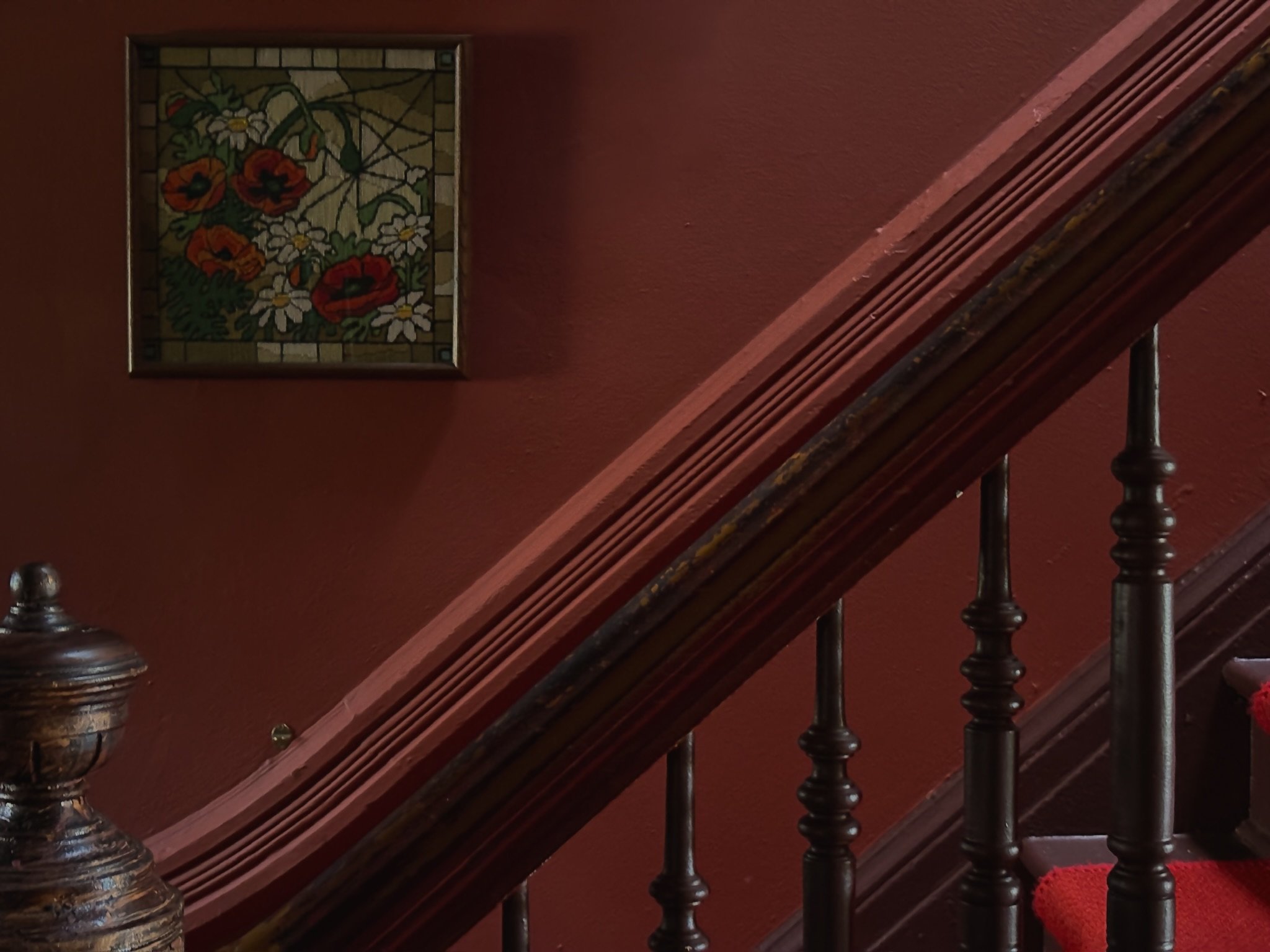 Close-up of a wooden staircase with red carpeting, and a stained glass floral window on a dark red wall.