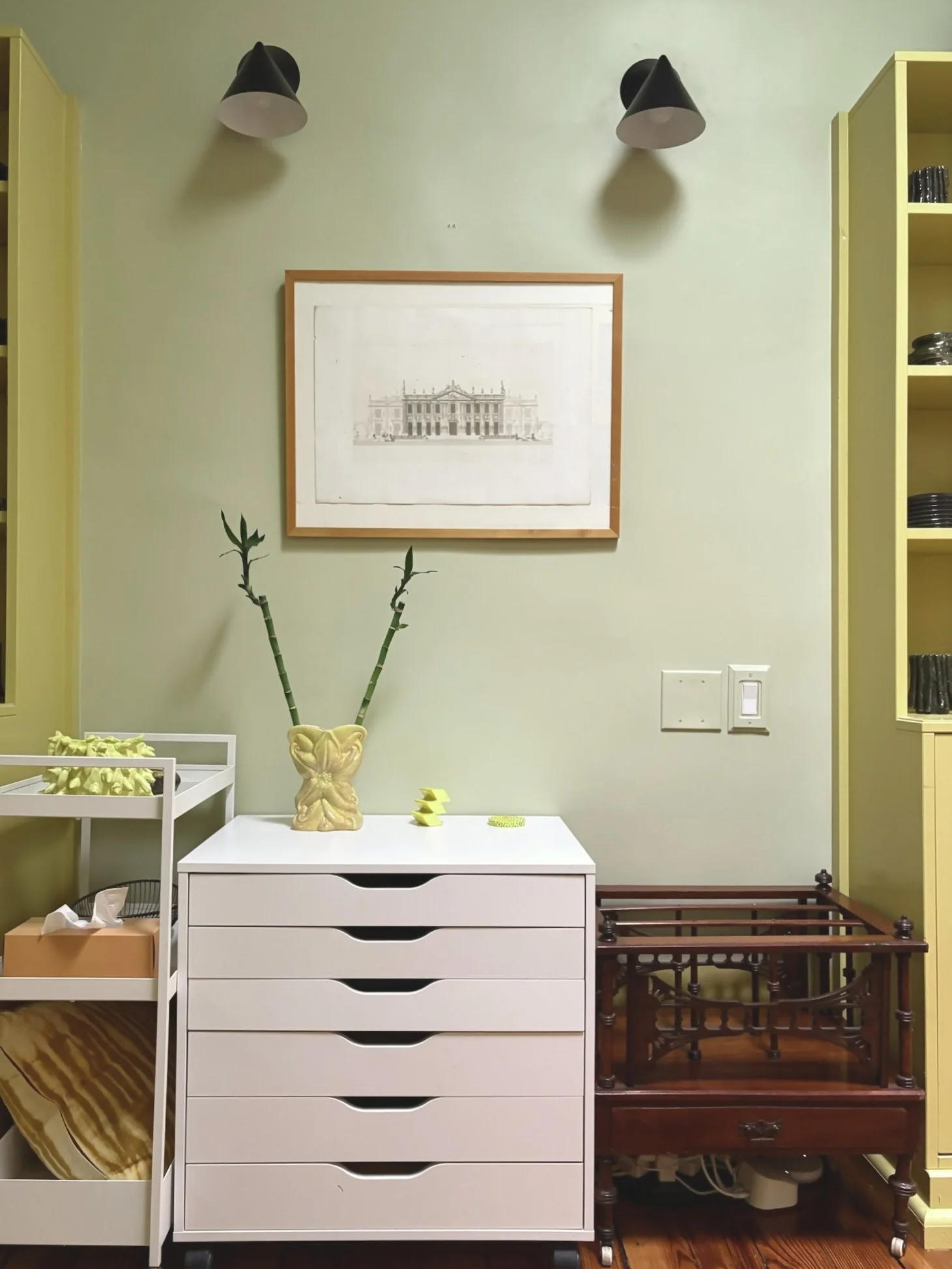 A room corner with a framed sketch of a building on a pale green wall, two black wall-mounted lamps above, a white dresser with handles, a tall yellow shelving unit on the right, and a small wooden table below the dresser. Decor items include a yello