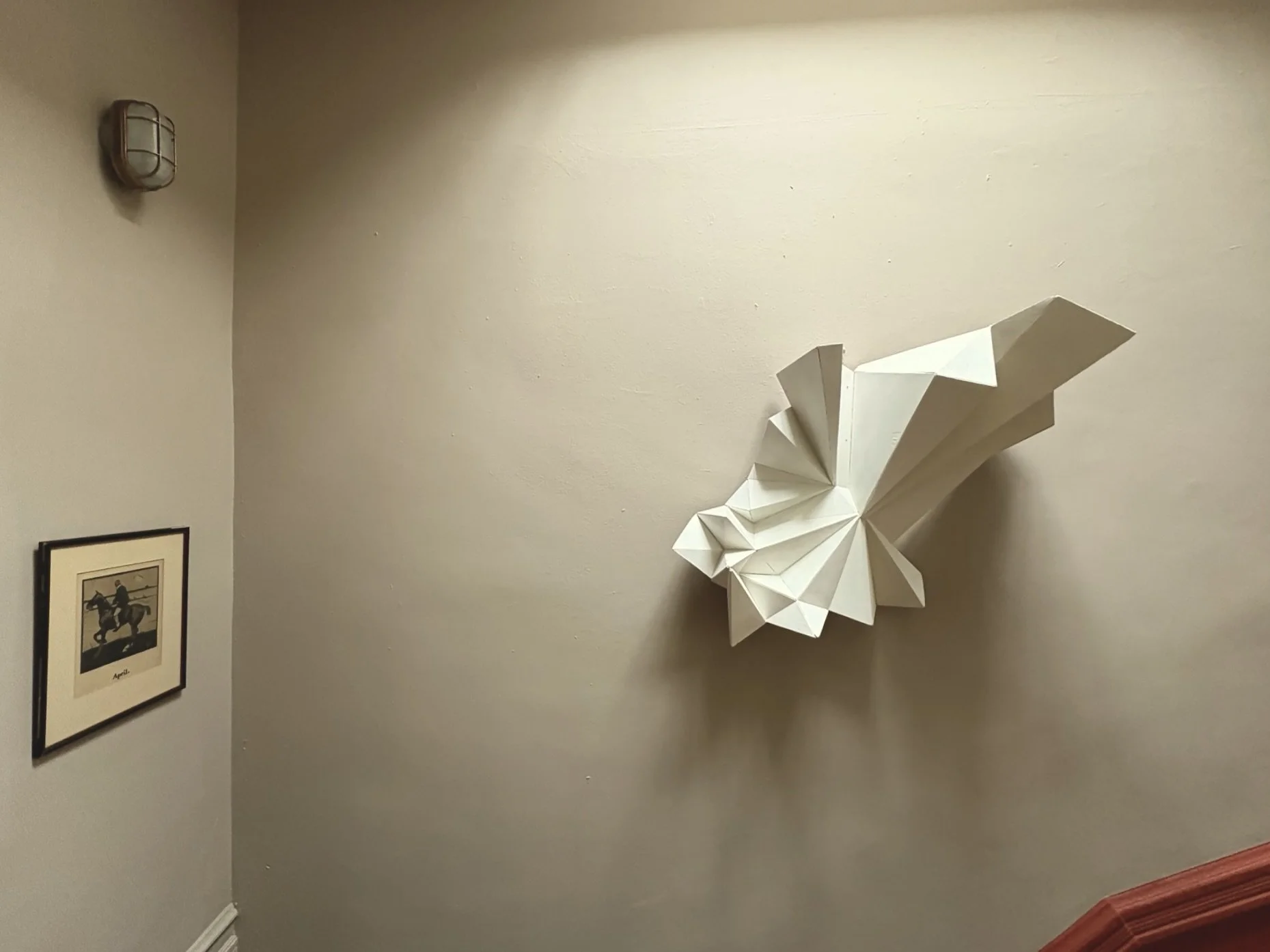 A beige wall with a white abstract paper sculpture resembling a butterfly or flower, a framed black and white picture of a person riding a horse, and a wall-mounted light fixture.