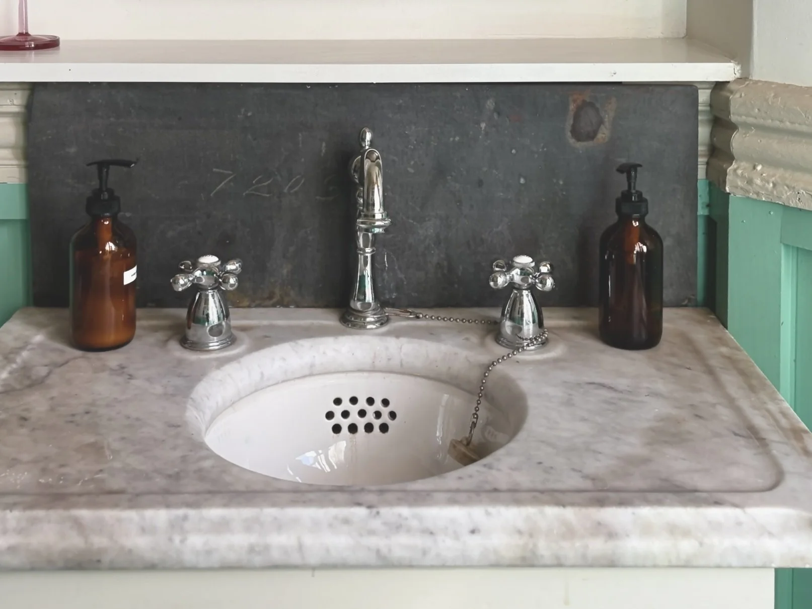 A vintage bathroom sink with a marble countertop, a chrome faucet, two amber soap dispensers, and a dark stone backsplash.