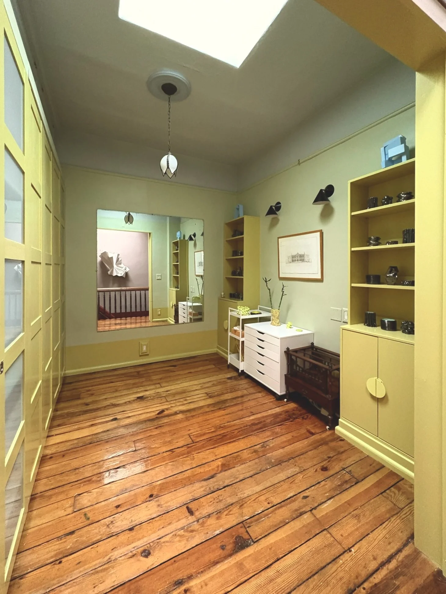 A room with yellow built-in shelves, hardwood floors, a mirror on the wall, and decorative lighting fixtures.