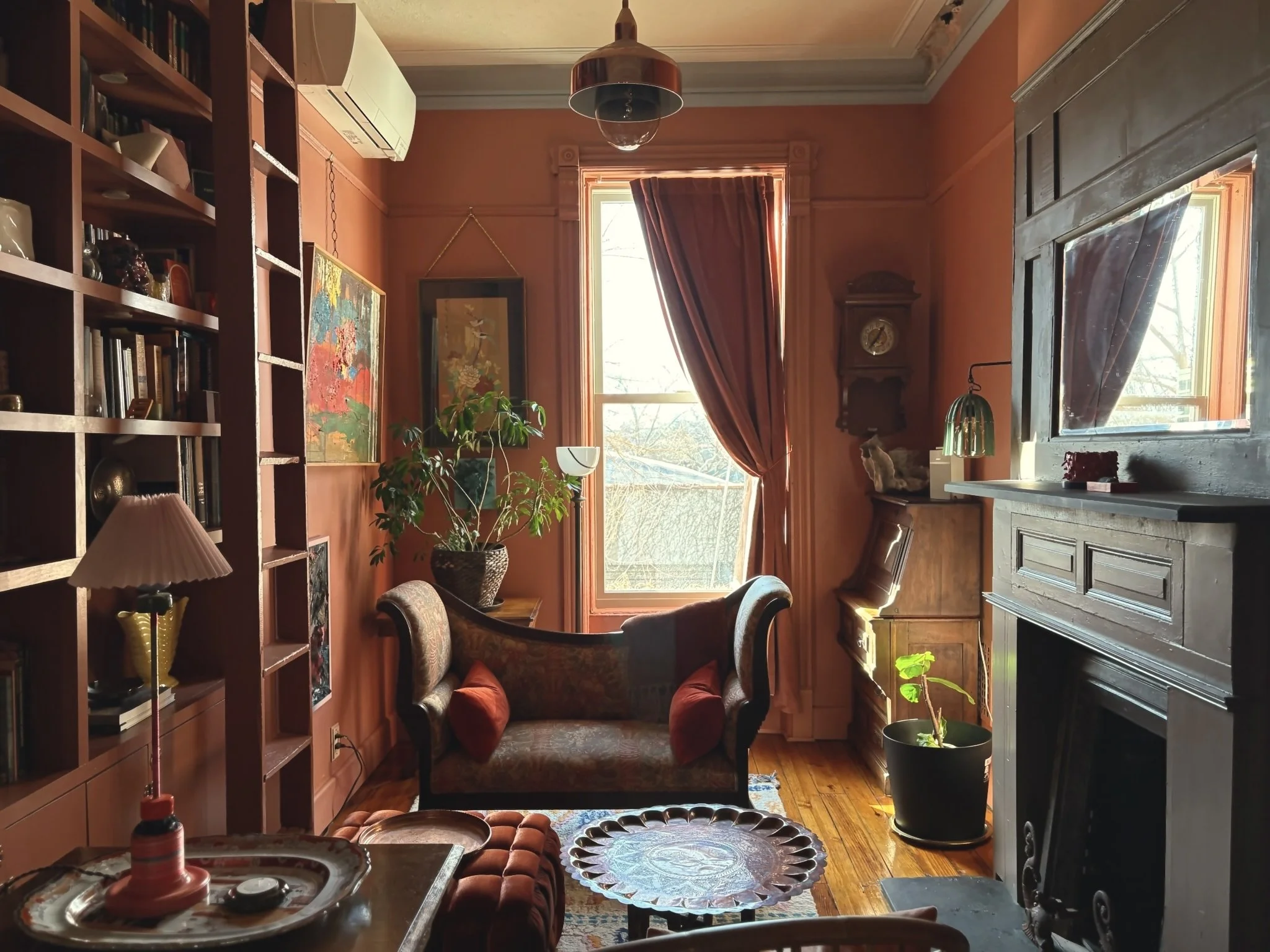 A cozy vintage living room with peach-colored walls, a large window with crimson curtains, an ornate bouquet, a dark wooden fireplace, a bookshelf filled with books, a framed painting, a small wooden cabinet, a potted plant, and various decorative it