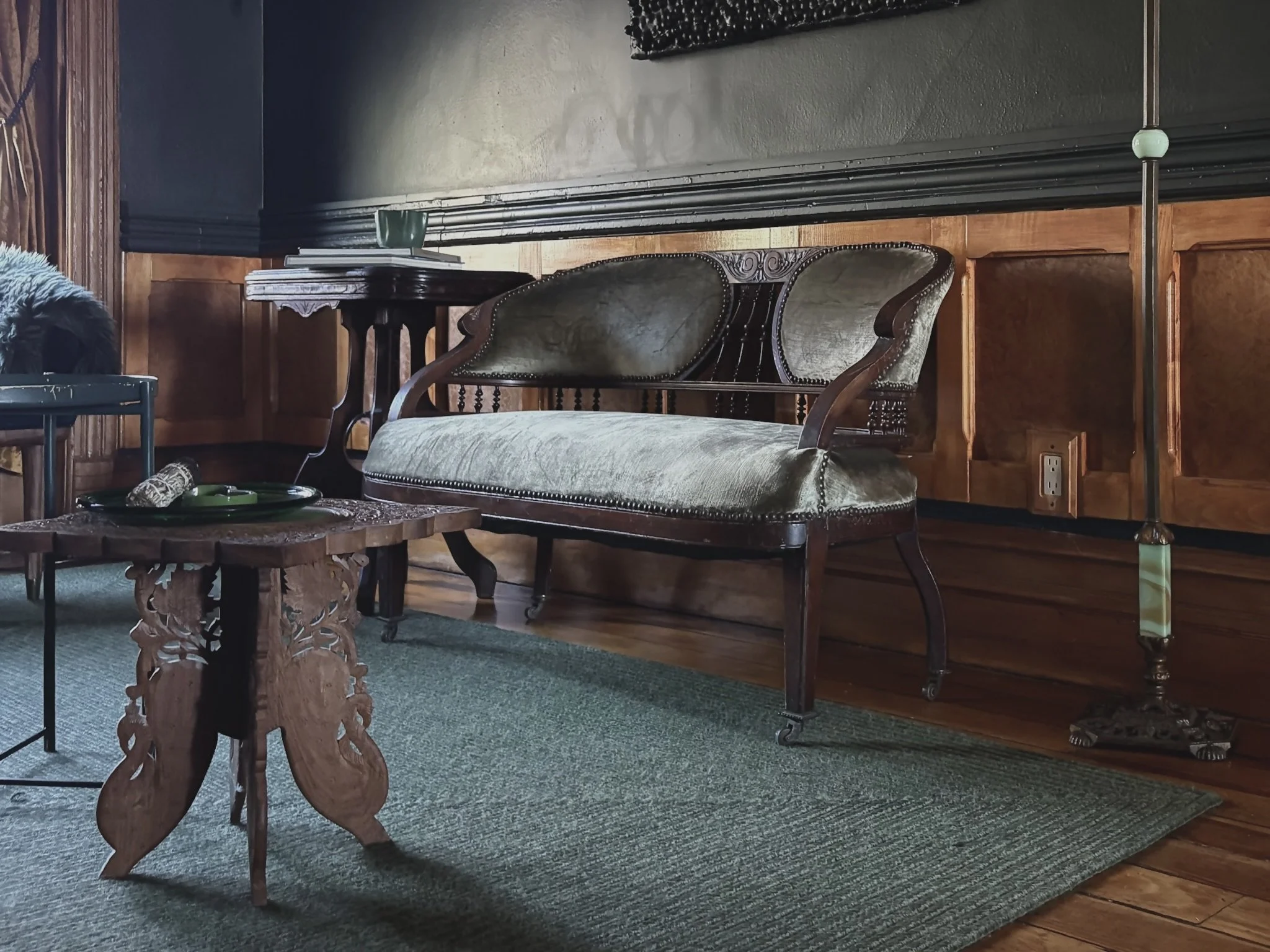 Vintage living room corner with ornate wood-paneled walls, a velvet-upholstered antique loveseat, a carved wooden side table, a small stack of books, and a lamp with a brass stand and patterned shade.
