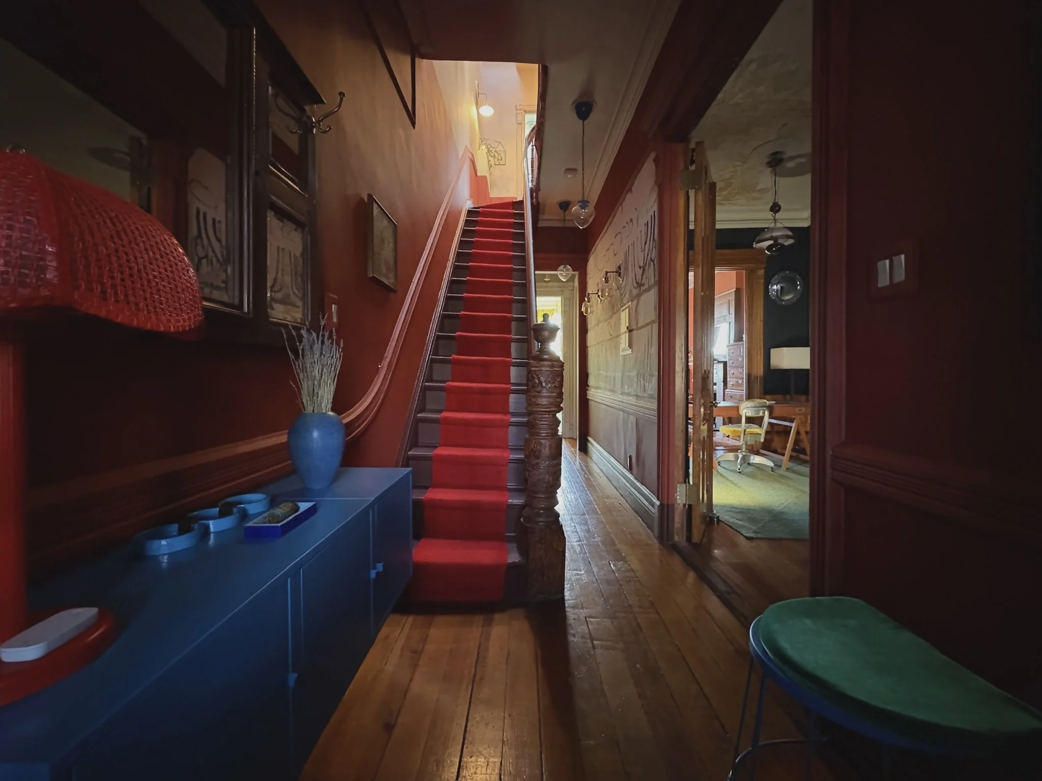 Entryway of a house with a wooden staircase, red carpet, and various decorative items, leading to other rooms with visible furniture and lighting fixtures.