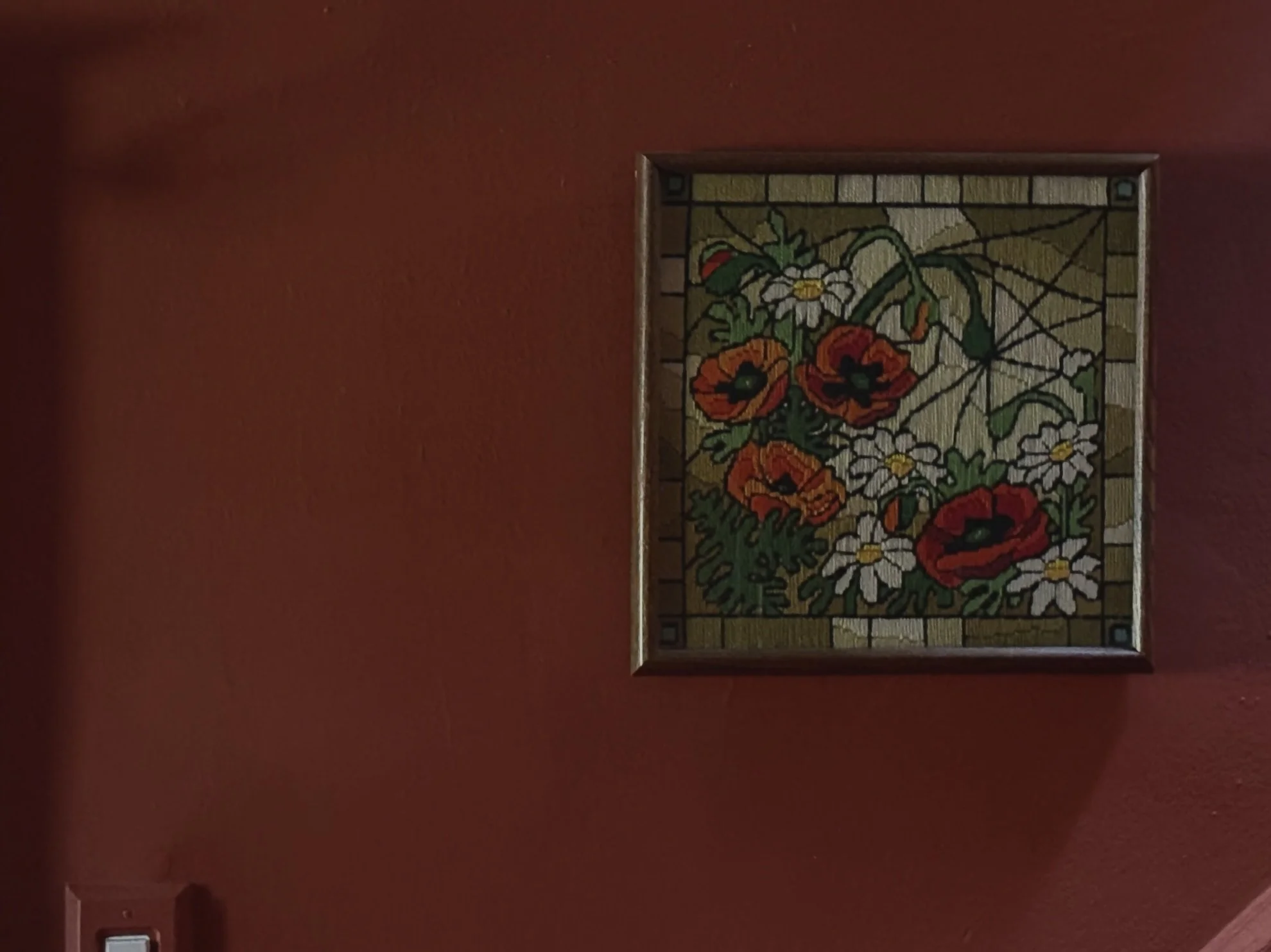 A framed colorful floral stained glass window featuring red poppies and white daisies, hanging on a reddish wall.