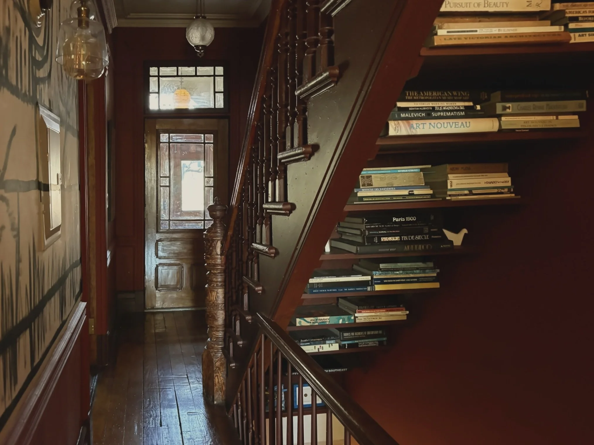 Interior view of a narrow hallway with a wooden staircase and a built-in bookshelf filled with books on the right side. The hallway has wooden flooring, a wooden door at the end, and patterned wallpaper on the left wall. Two hanging light fixtures ar
