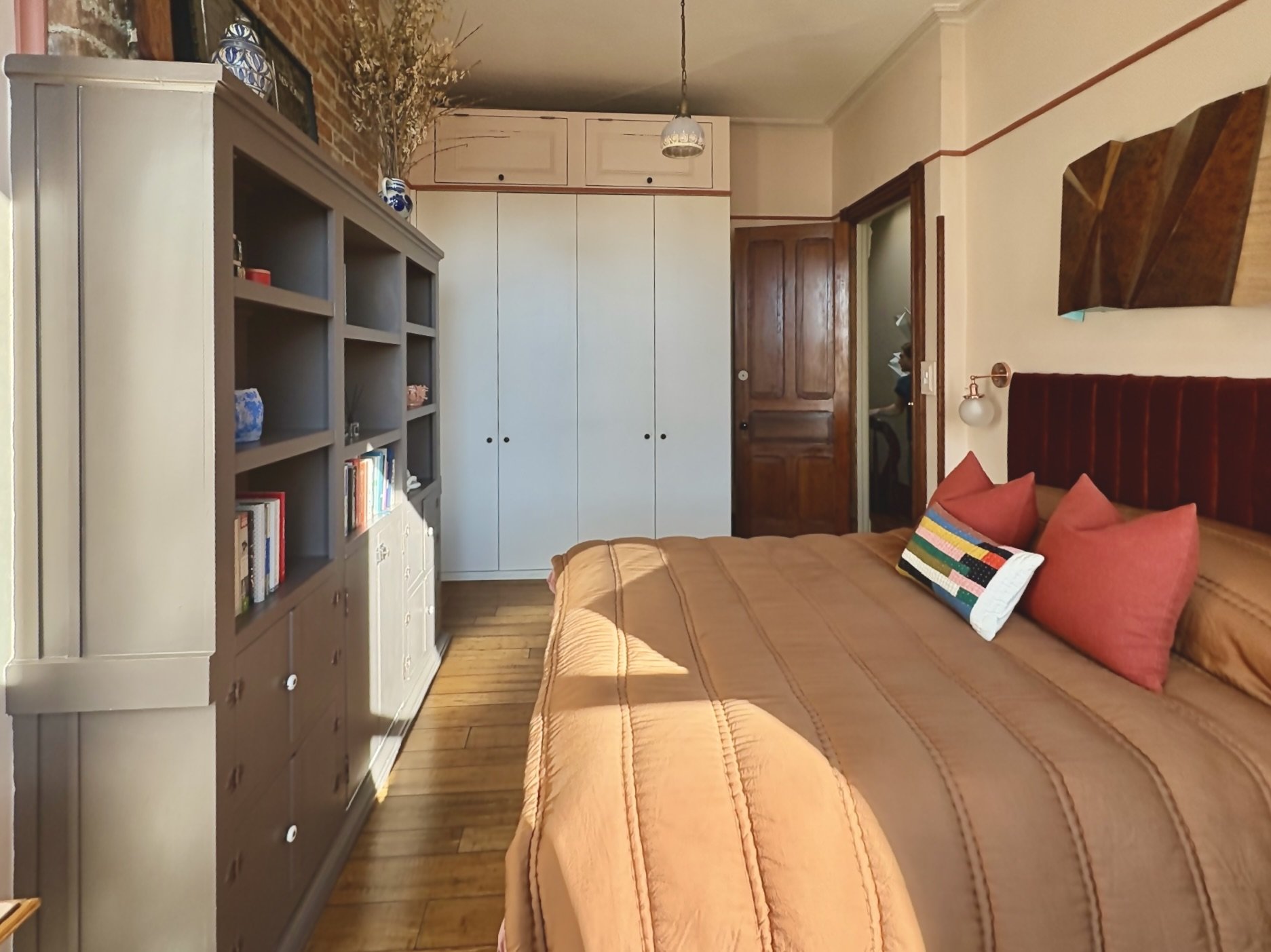 Bedroom with a bed, pillows, a bookshelf, a wardrobe, and a door, with wood flooring and a hanging light fixture.