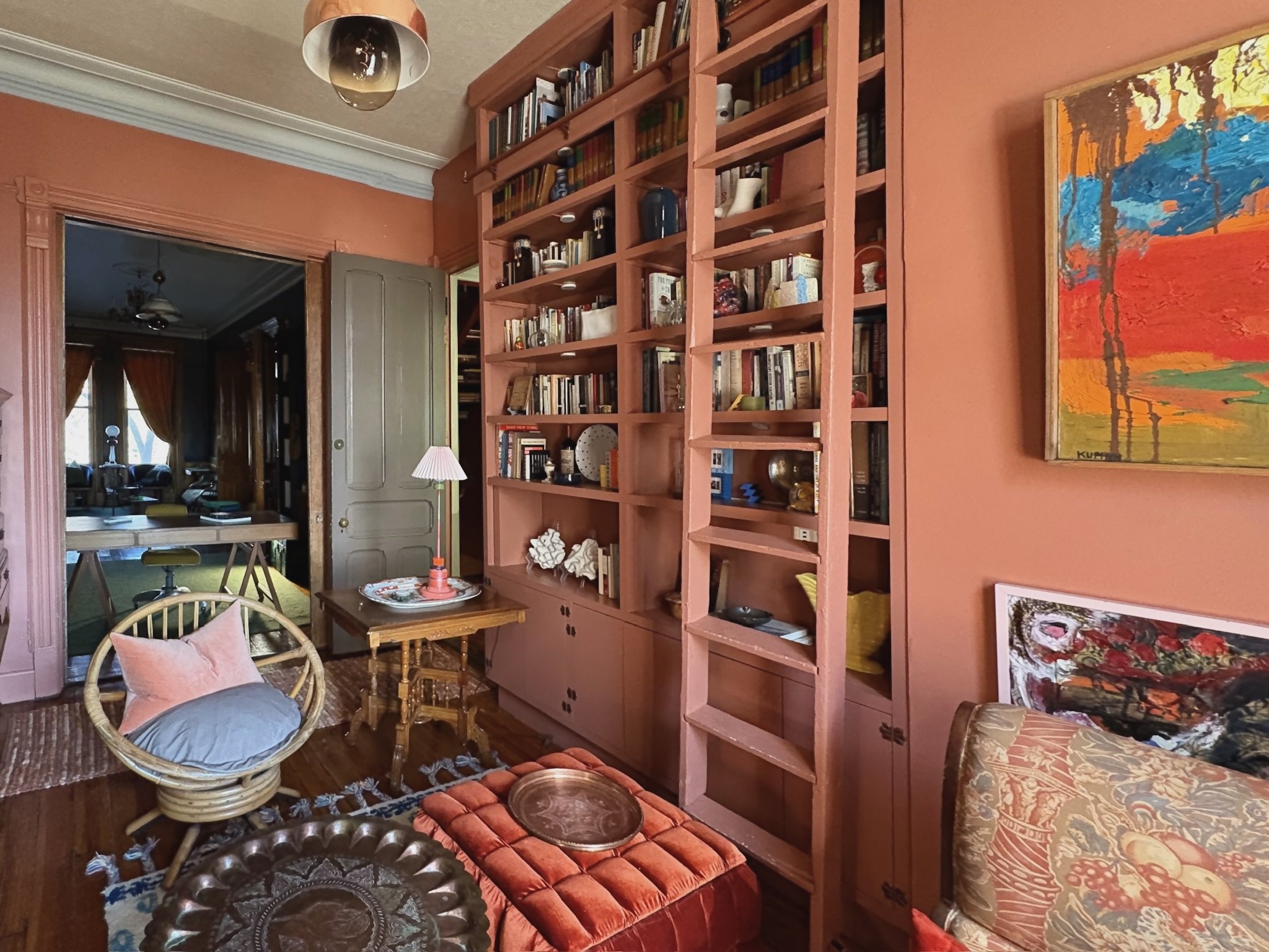 A cozy living room with pink walls and a built-in bookshelf filled with books and decorative items. There is a wooden table with a small lamp and decorative objects, a rattan chair with cushions, and a colorful abstract painting hanging on the wall. 